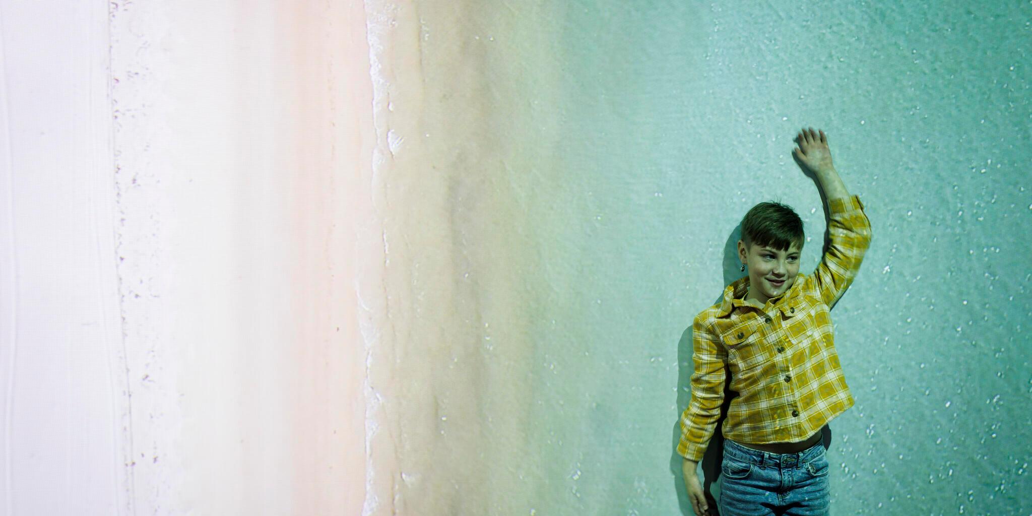 Young boy with short, dark hair, wearing a yellow check shirt standing in front of a large projection of a coastline