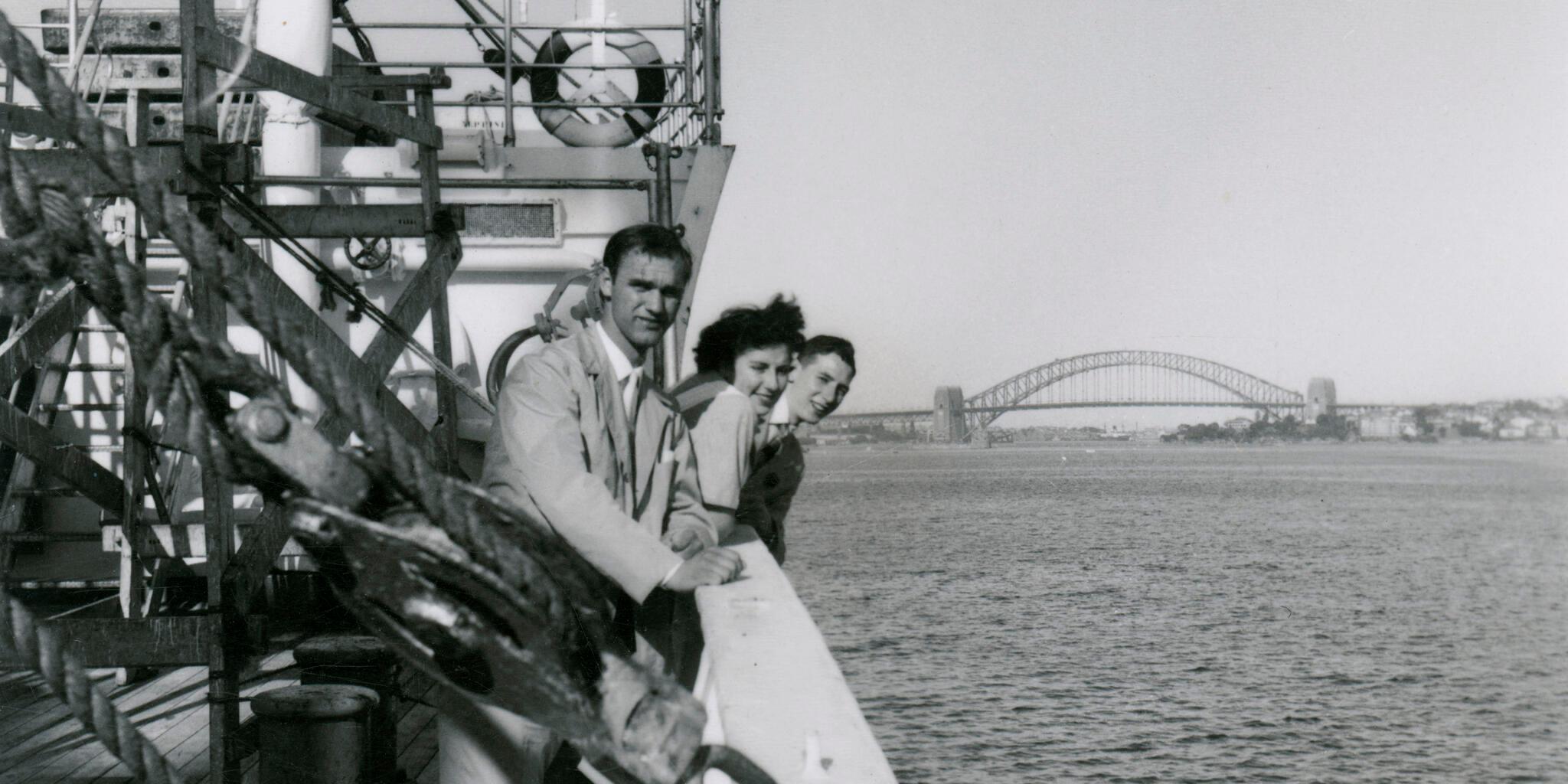 Black and White photograph, three passengers on the deck of migrant ship MV NAPOLI as it moves up Sydney Harbour. The Harbour Bridge can be seen in the background.