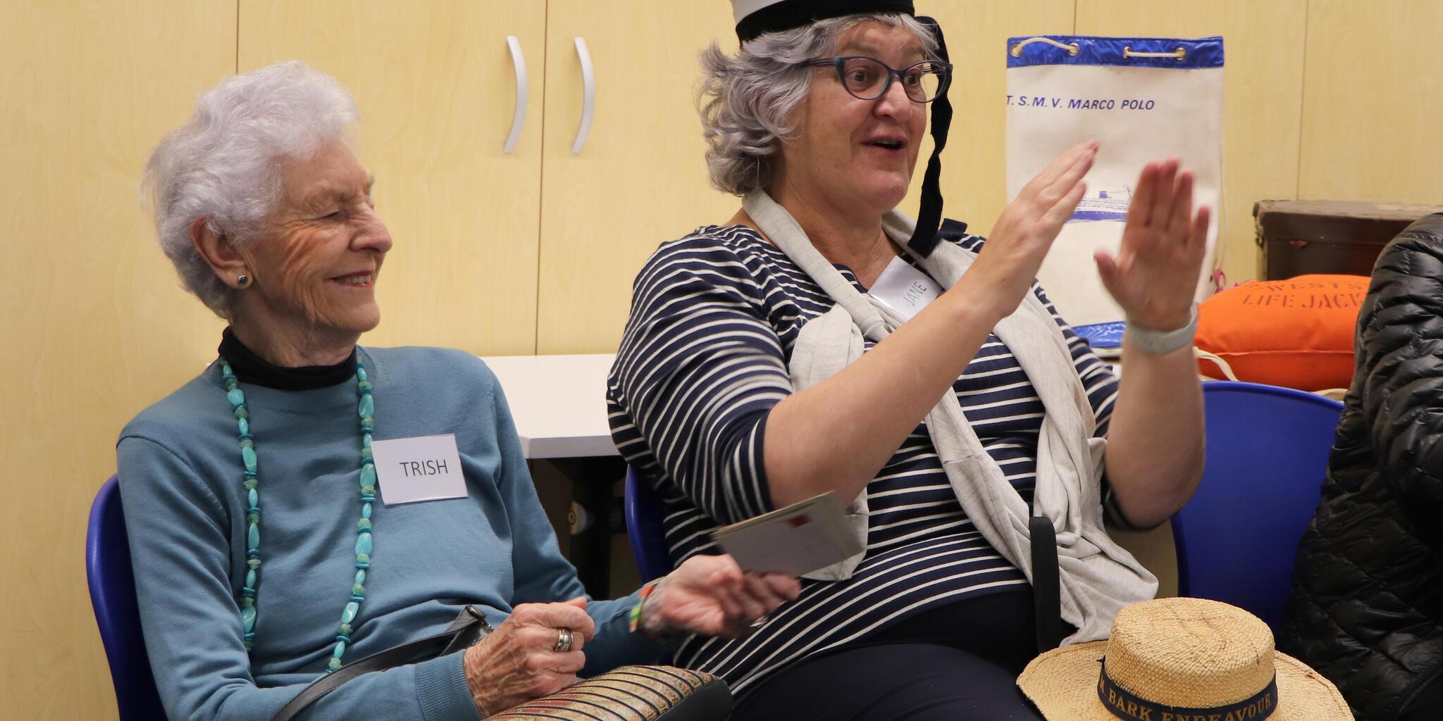 A lady with short white hair and a blue jumber sitting next to another lady with a stripped top and sailor's hat. 