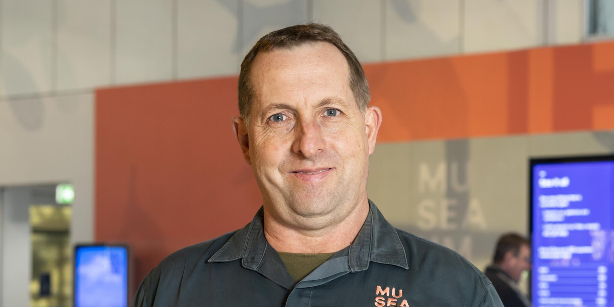 close up photo of a man with short hair and white skin, wearing a museum volunteer uniform with military service medal ribbons 