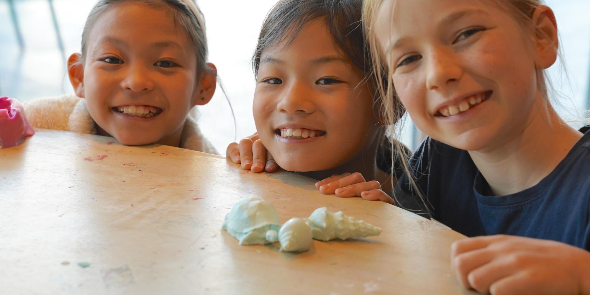 3 girls faces on the edge of a table, with some shell artworks.