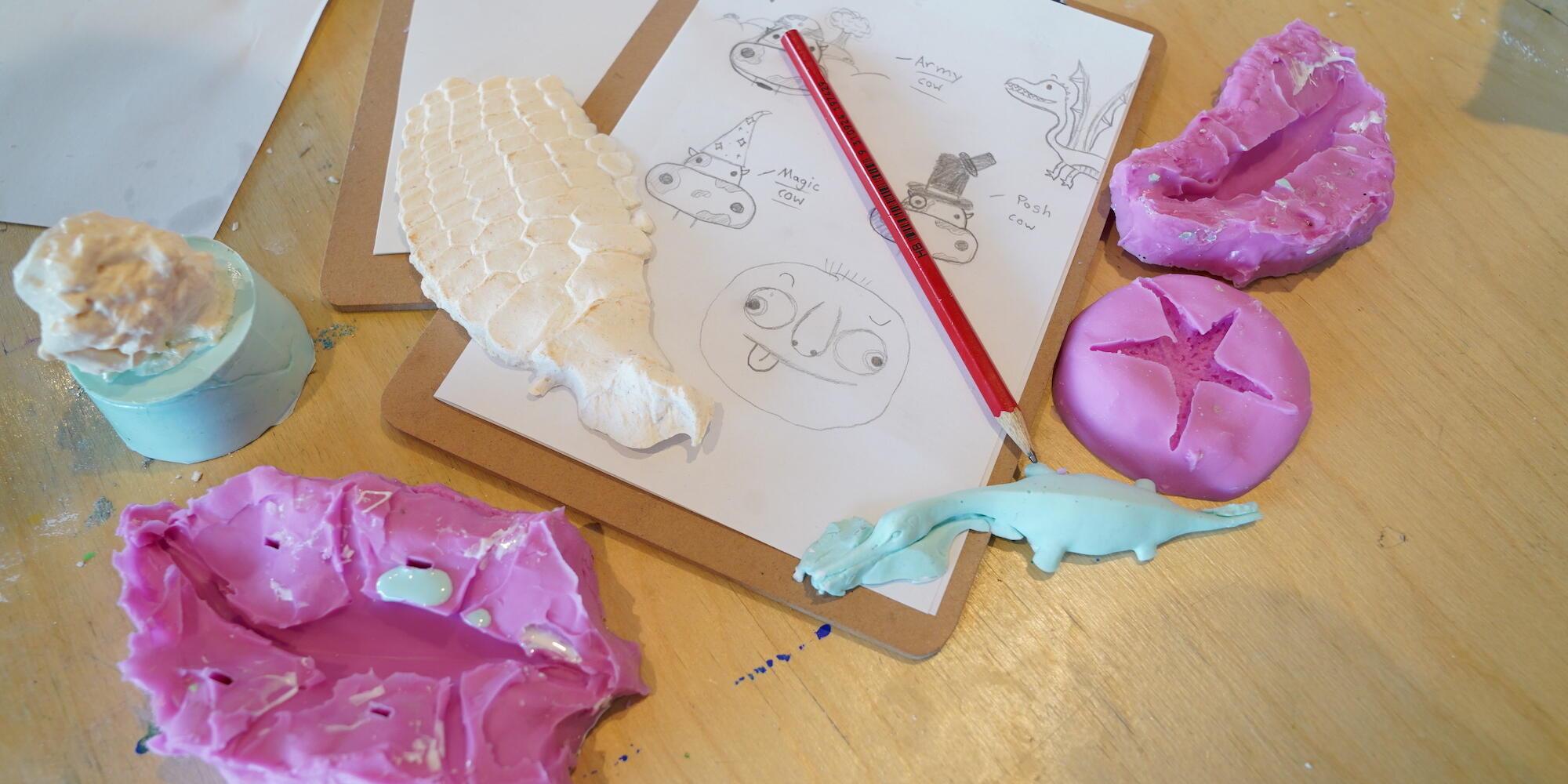 A table with a clipboard with paper containing pencil drawings, surrounded by pink silicone moulds 