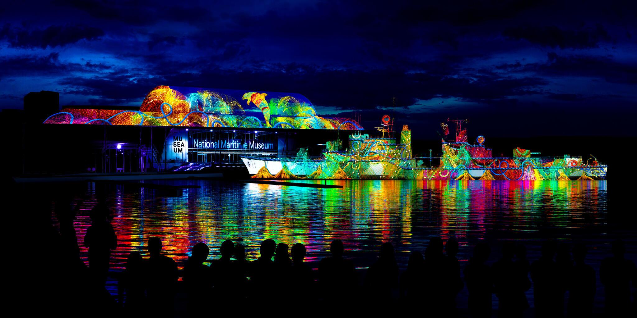 photo of colourful projection on the side of the museum and navy destroyer at night, with multicoloured reflections on the water. 