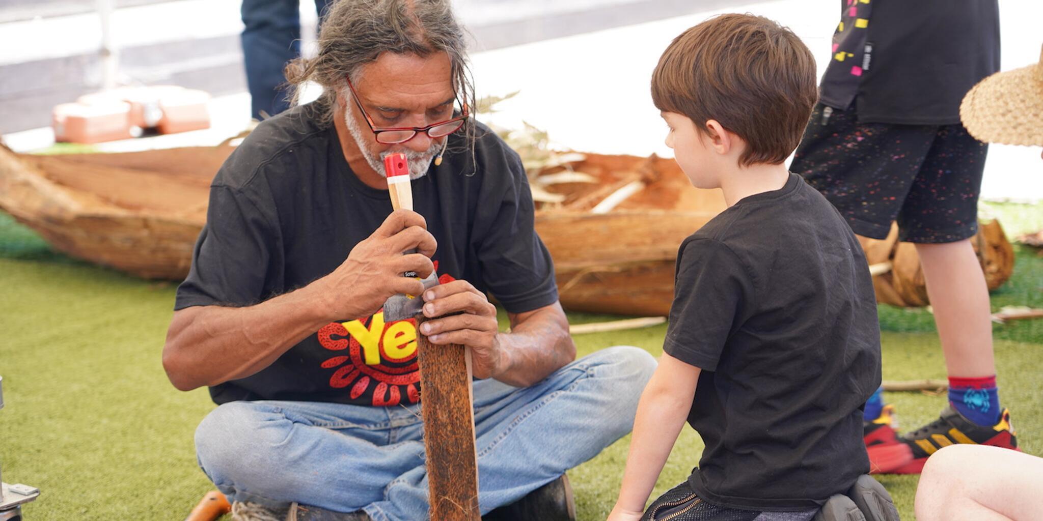 Man and a boy kneeling on the ground. The man us using a tool to work on a piece of bark.