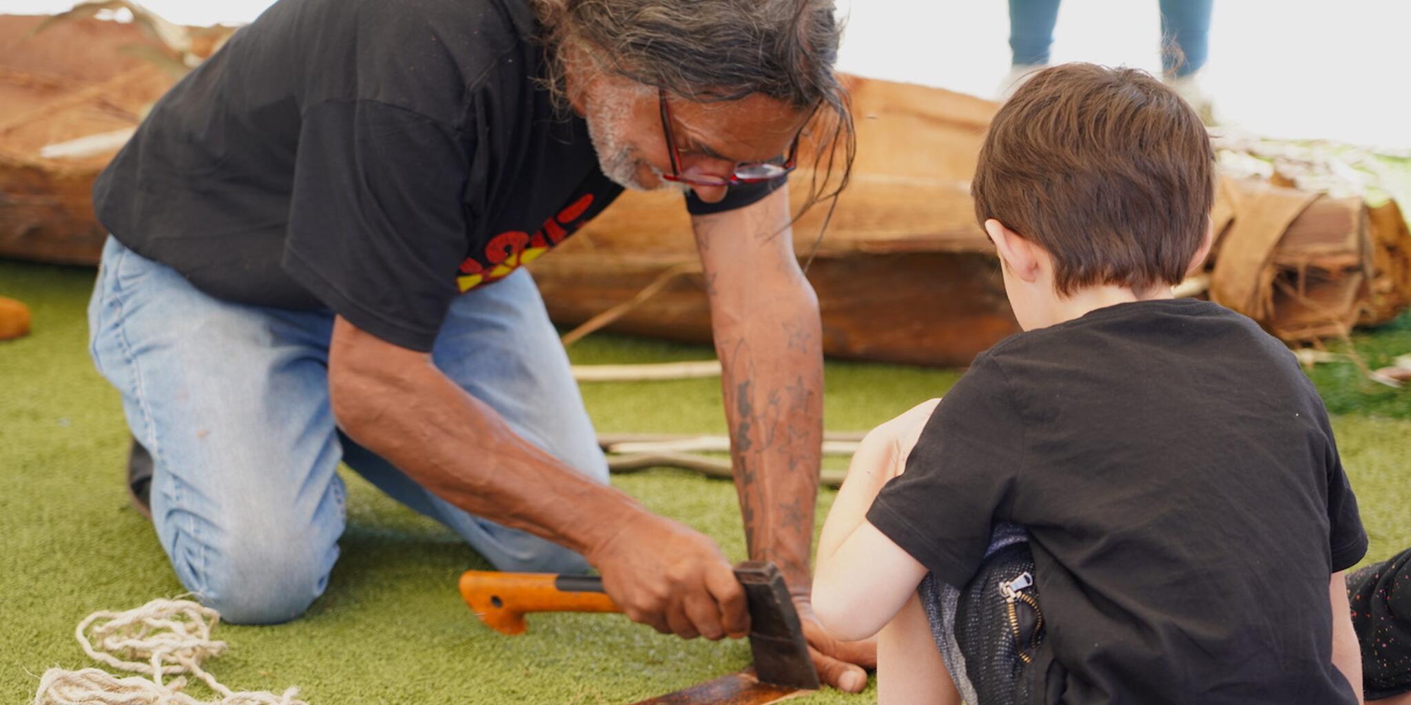 Man and a boy kneeling on the ground. The man us using a small axe to work on a piece of bark.