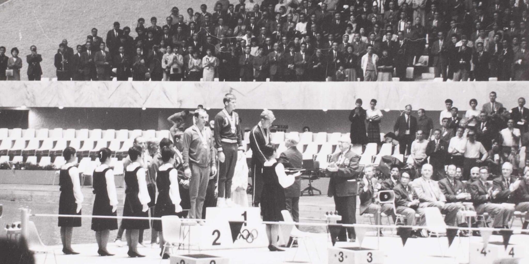 Black and white photo showing a medal podium with people standing on it and a crowd behind them.