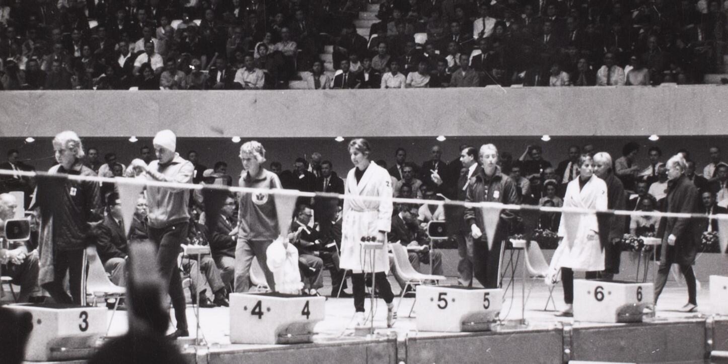 Black and White photo showing a swimmers walking out to one end of a pool preparing for a race with a crown behind them.