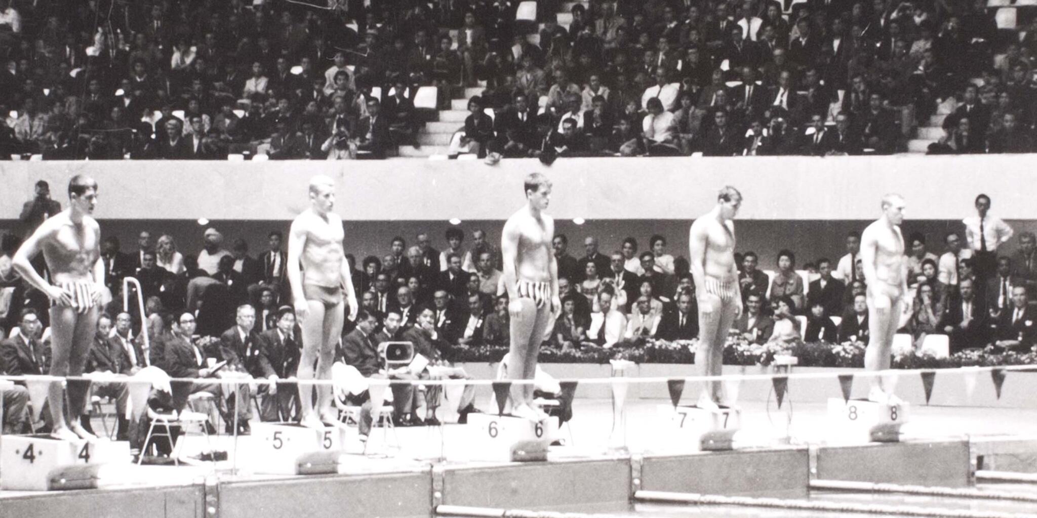 Black and White photo showing a line of 5 swimmers at one end of a pool preparing for a race with a crown behind them.