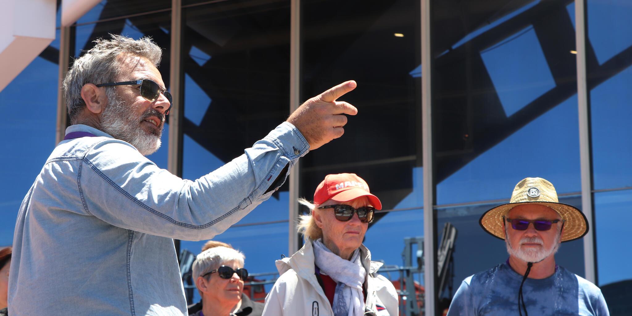 A man with grey hair and his back to the camera is pointing to the right. There are other people in the background wearing hats and sunglasses. 