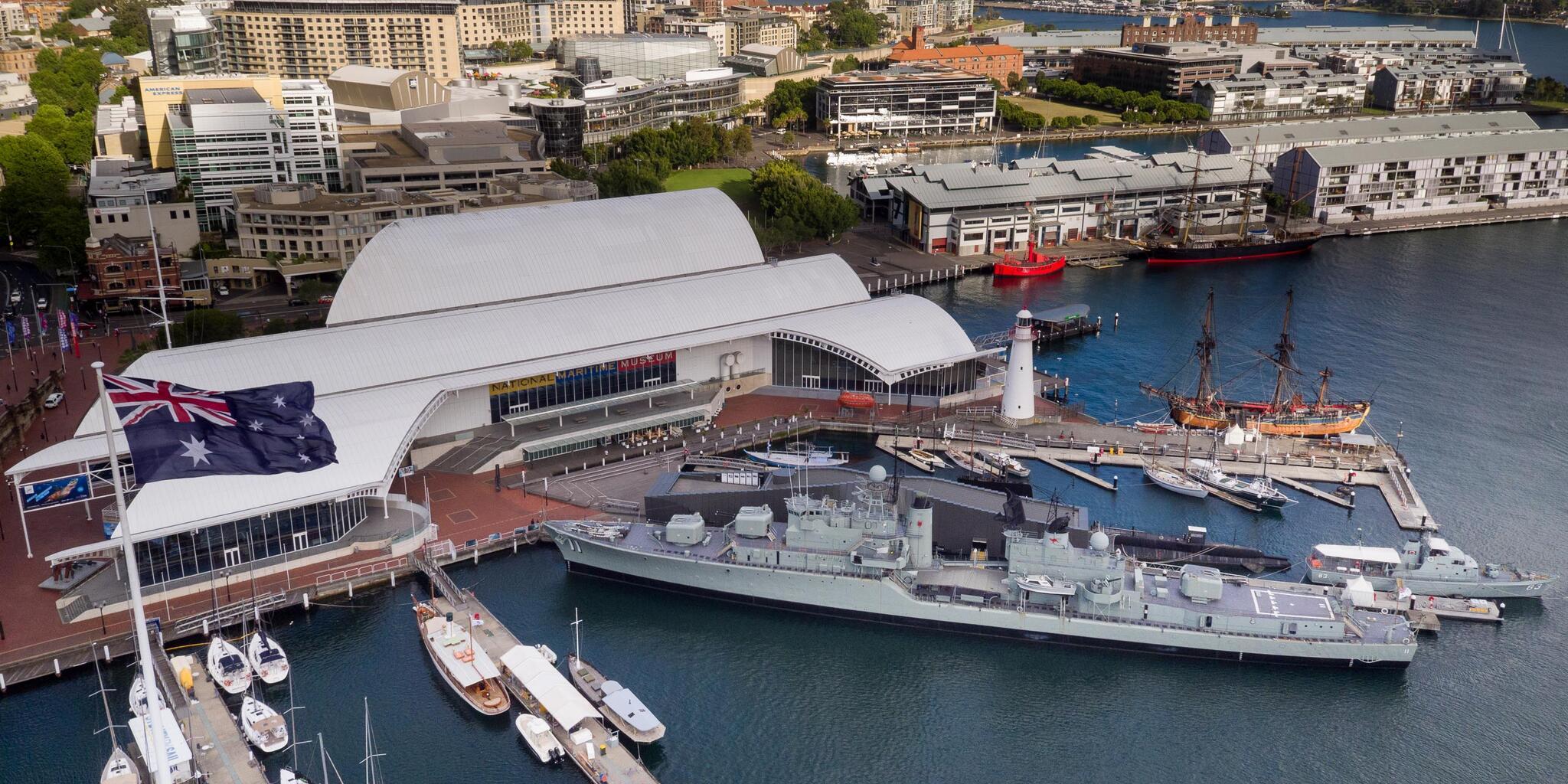 Aerial view of the museum shot by drone. Museum building at centre with Wharf 7 building at upper right. There is an Australian flag, and various wharves and museum vessels. 