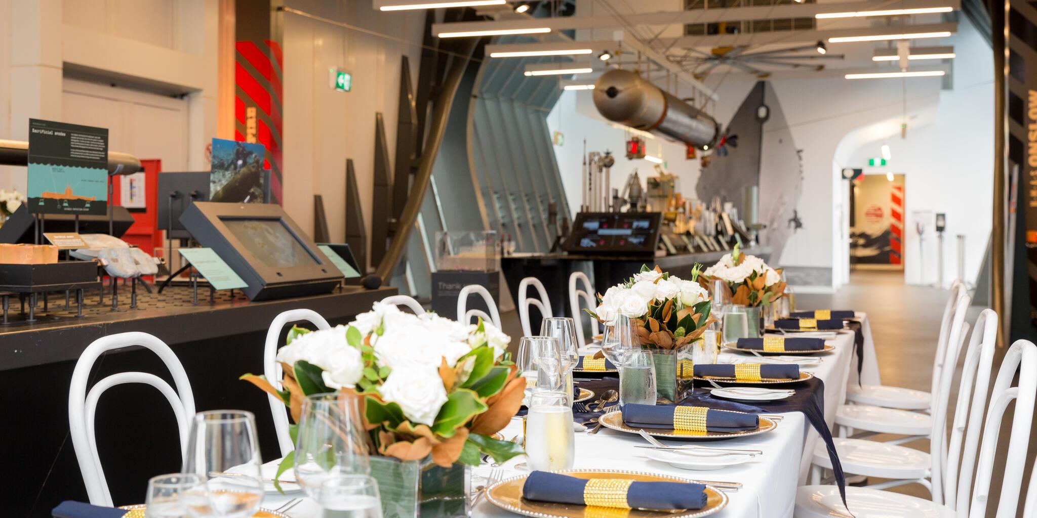 photo of a long table with white tablecloth set for a formal event and decorated with flowers, in a museum space.