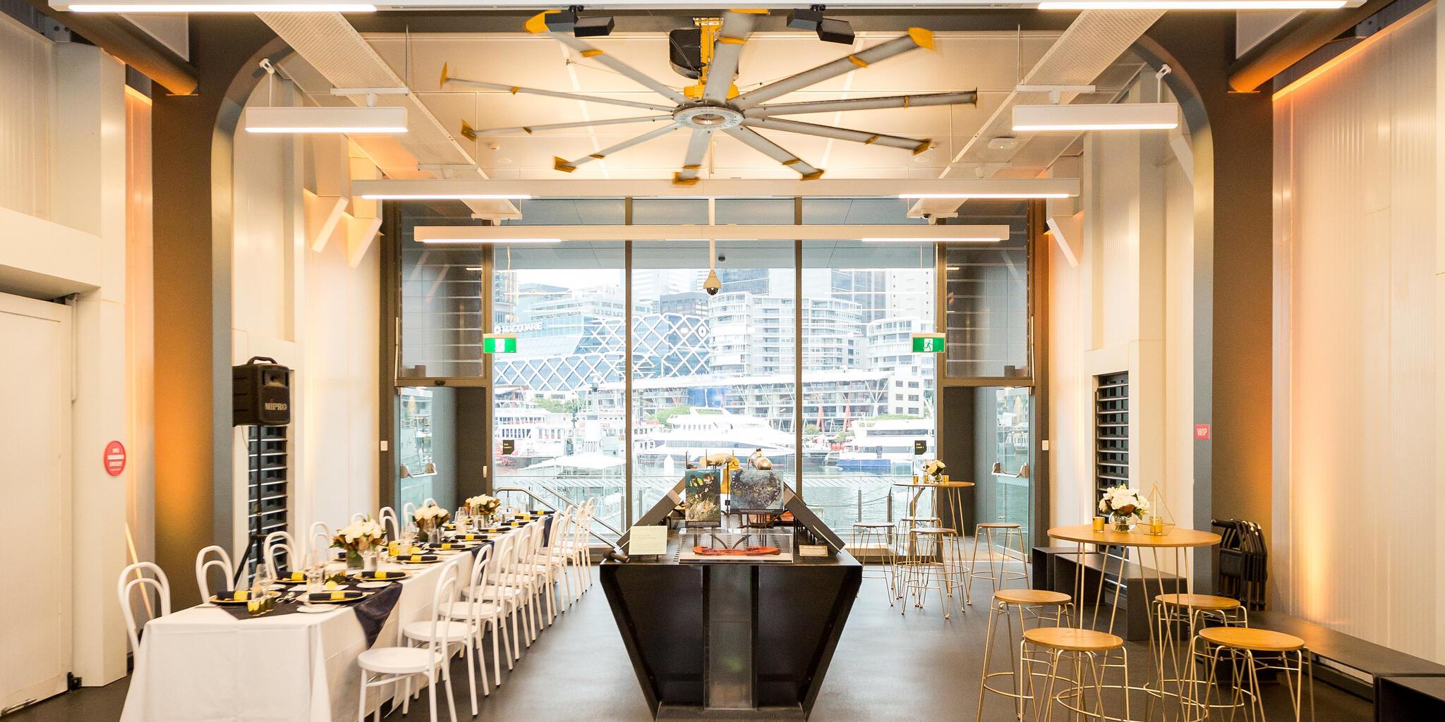 photo of a museum space with a long table set and decorated on the left side, museum interactive displays in the middle and a cocktail table and stools on the right, with a large window to the rear.  