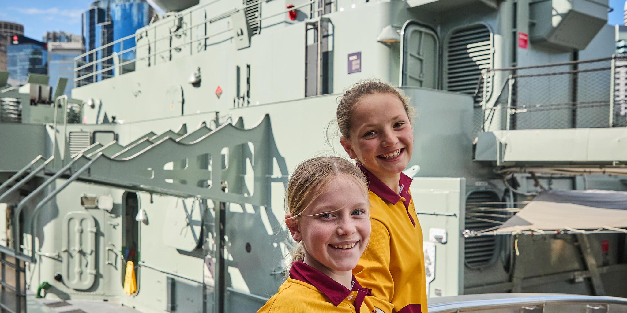 Two firls in a yellow and red school uniform on a gangway with the navy vessel vampire behind them.