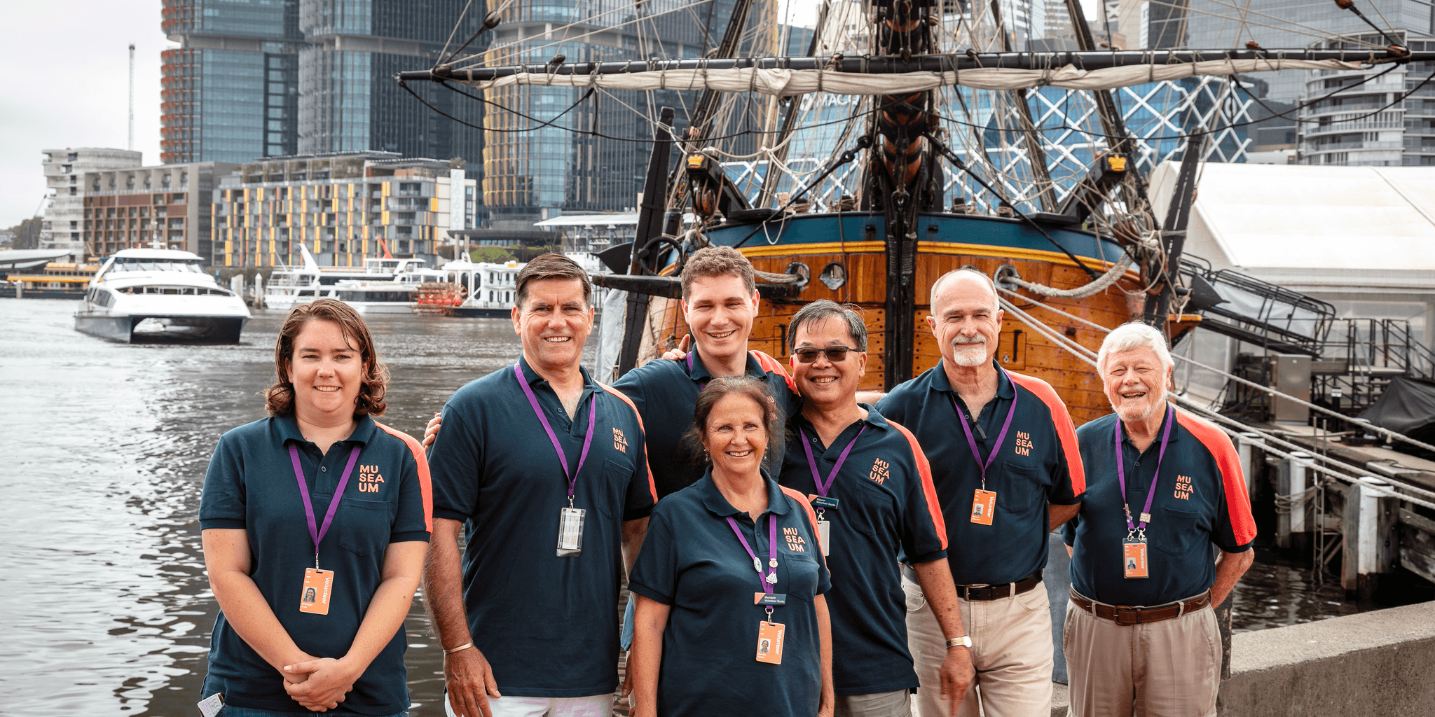 6 volunteers in navy museum unforms posing with HMB ENDEAVOUR replica tall ship in background.