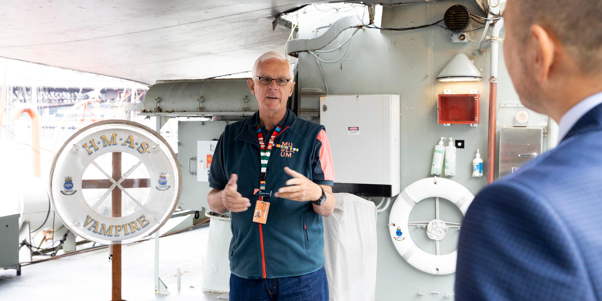 Photo of a male in a navy volunteers uniform standing on the deck of navy ship, the the shoulder of a man in the audience on the right side of the image.