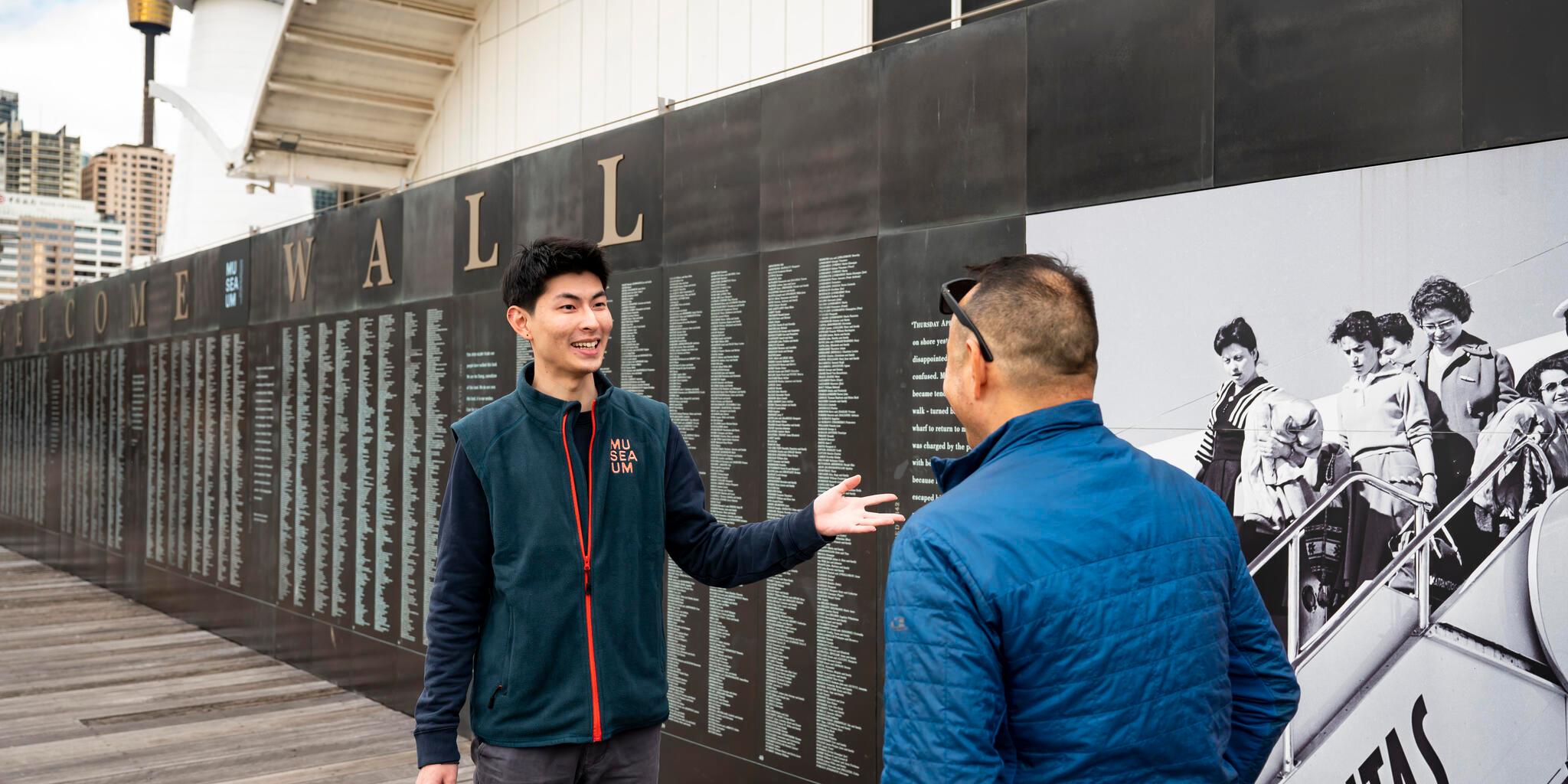 Male volunteer in a uniform showing a visitor names engraved on the national monument to migration