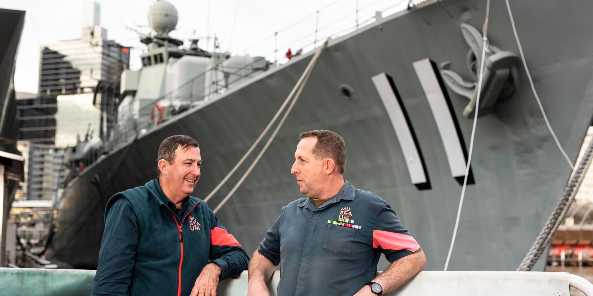 2 men in volunteer uniforms with the navy destroyer VAMPIRE in the background