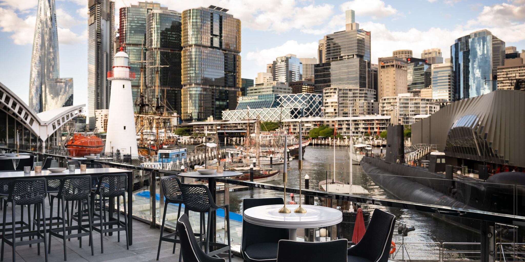 Photograph taken on a sunny day with tables and chairs in the foreground, with a lighthouse and the Sydney skyline in the background