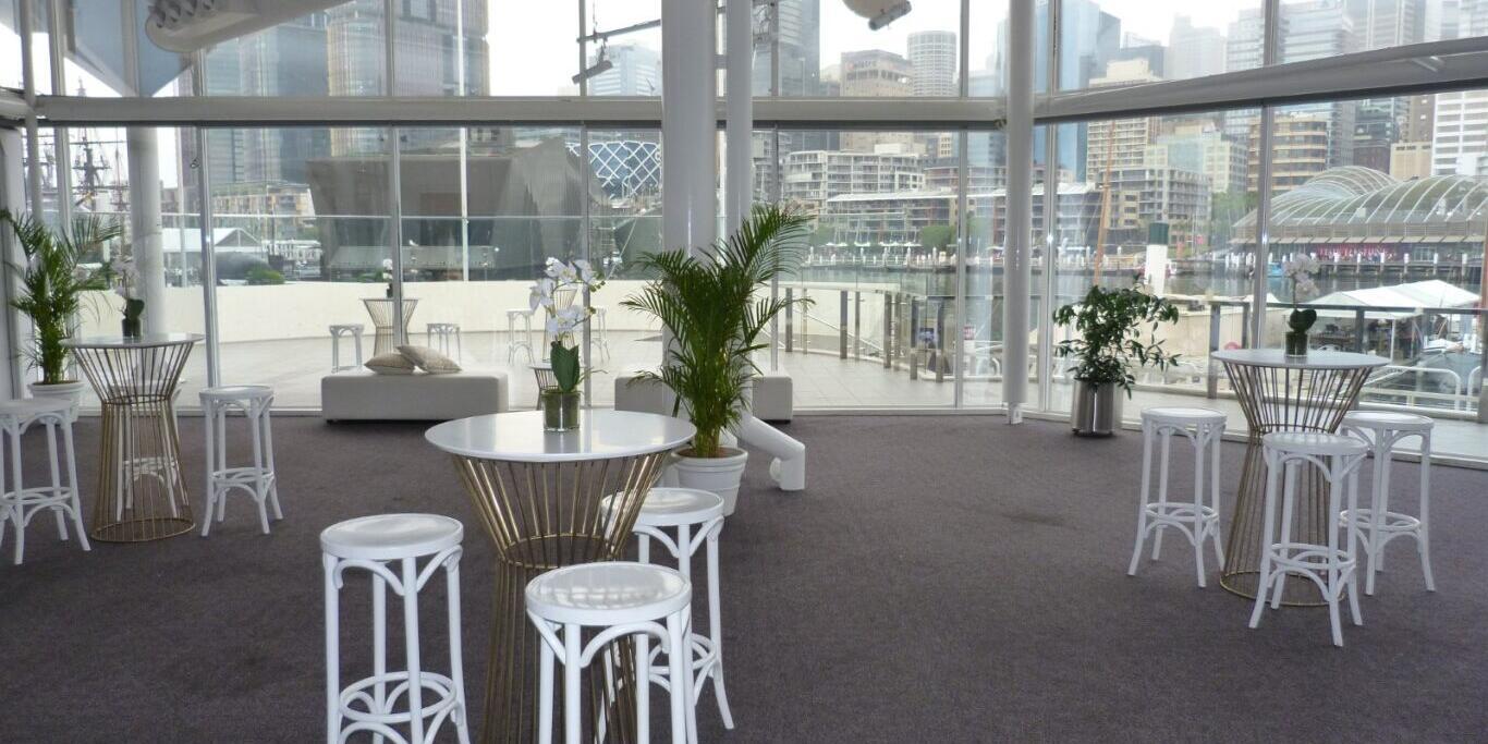 photo of an interior space with cocktail tables, stools and plants, with a full hight windows in the background.