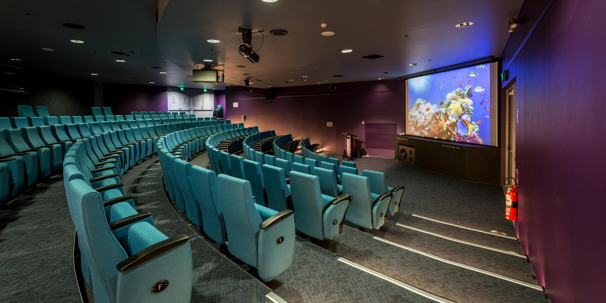 photo of an empty theatre with blue seats and a large screen at the front. 