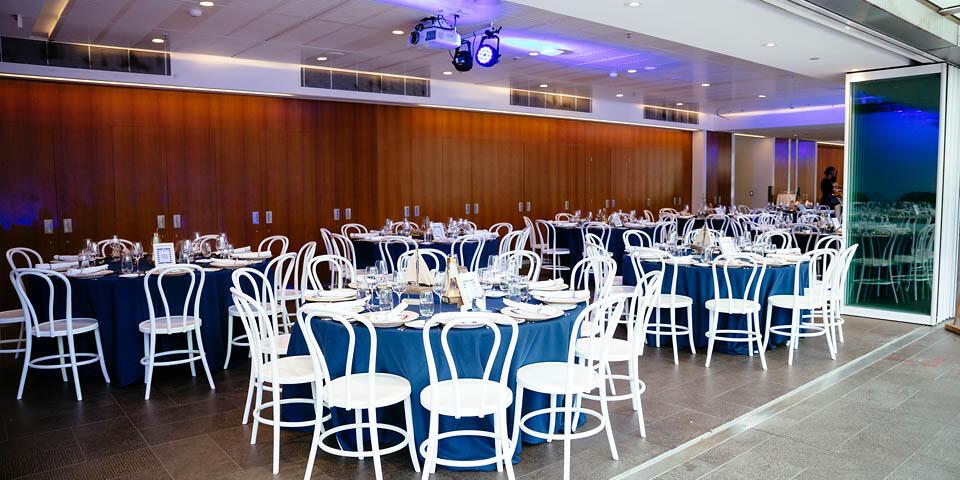 photo of event space with white chairs and large round tables with blue table cloths and place settings. 