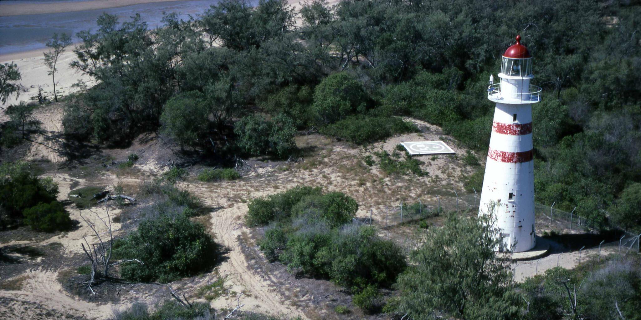 Aerial photograph showing the lighthouse on the right surrounded by scrub with beach visible at upper left.