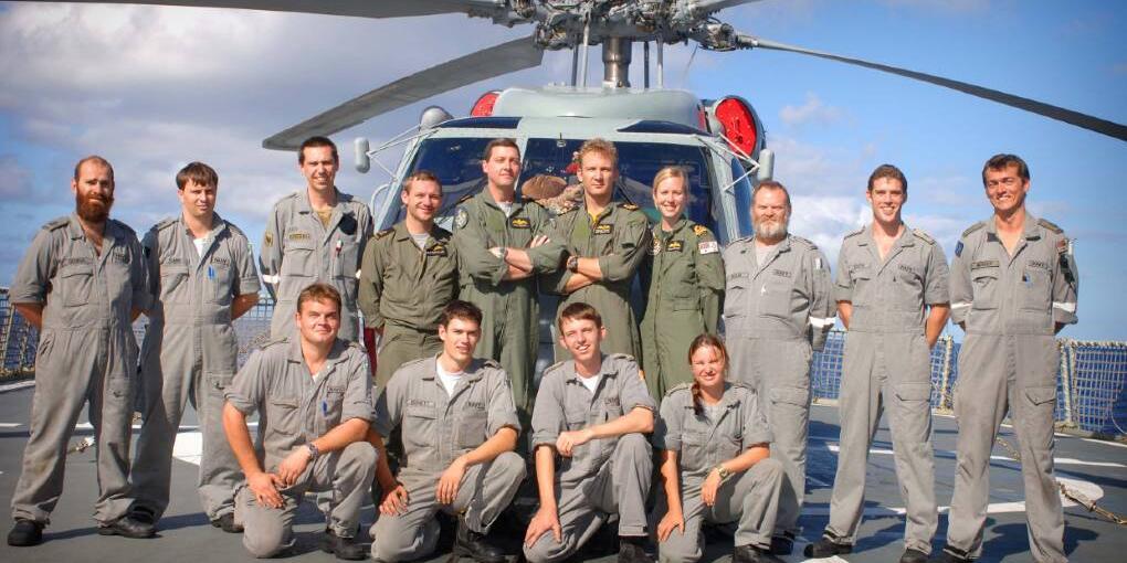 A group of military personel in uniform. The photo is taken outside standingg or kneeling infront of a grey navy helicopter. 