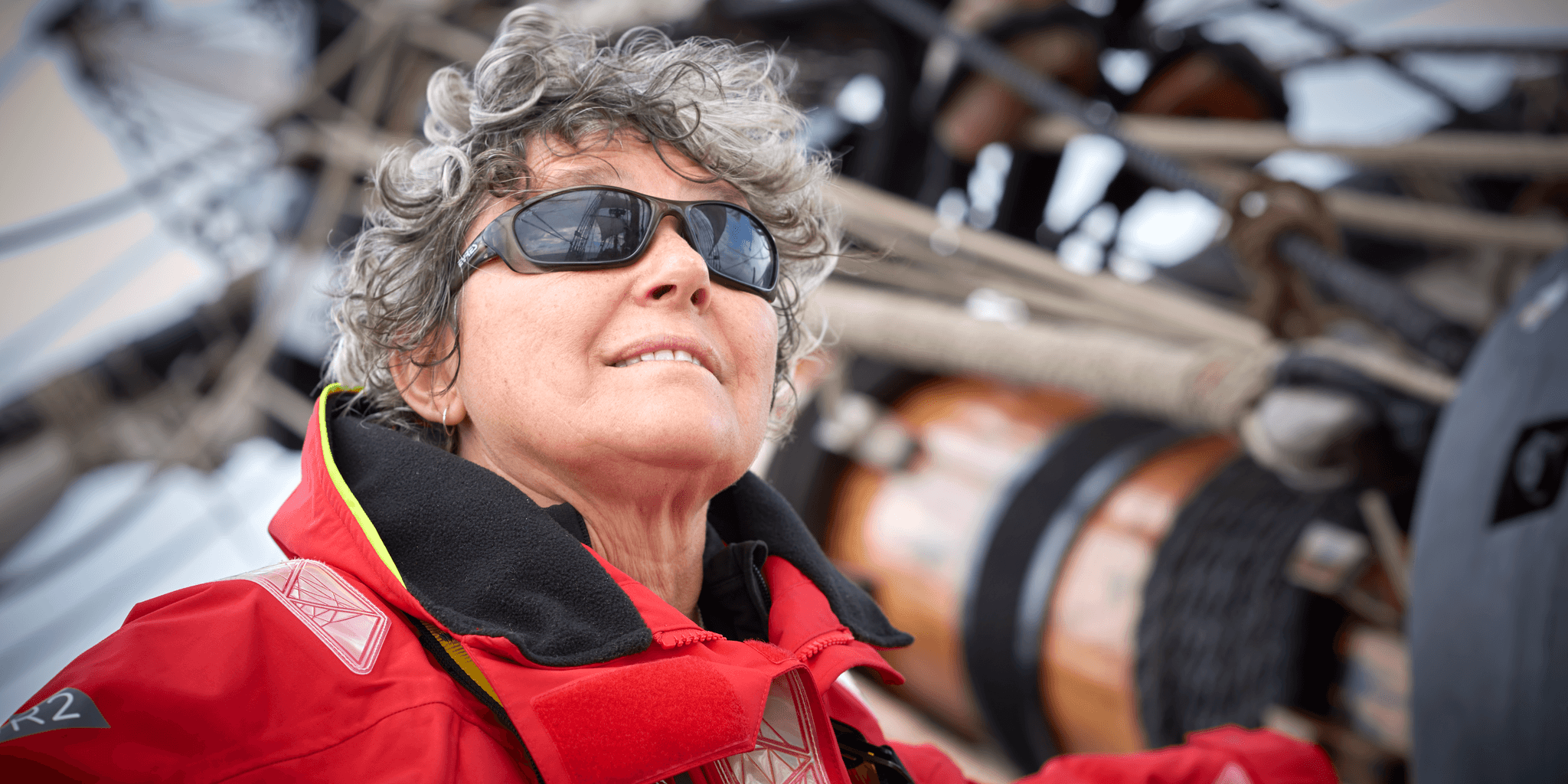 Above deck, woman wearing sunglasses looking out to sea with mast in background. 