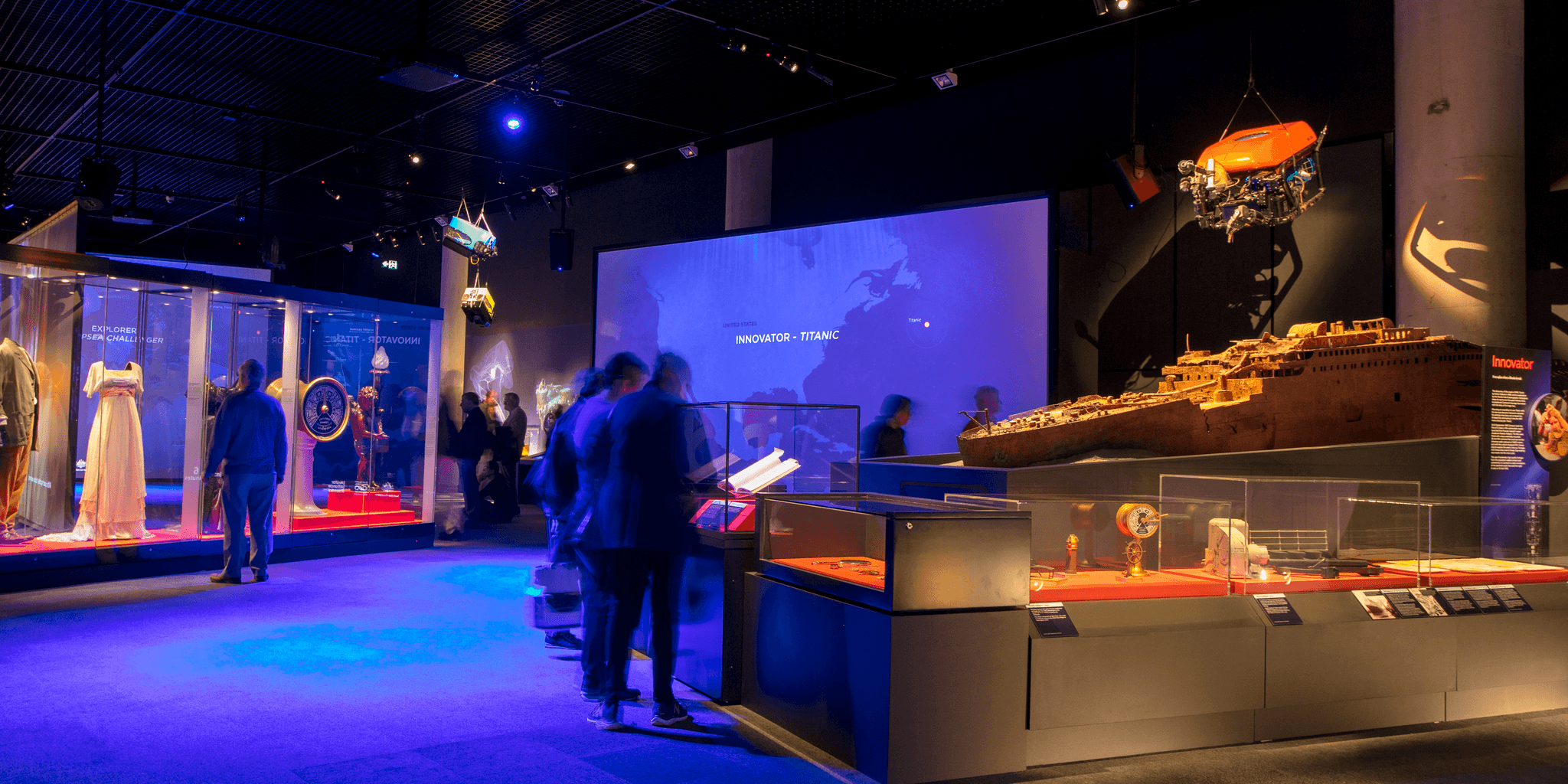 Photo of an exhibition space with blury figures lookiing at a display case with costumes and a large model ship.  