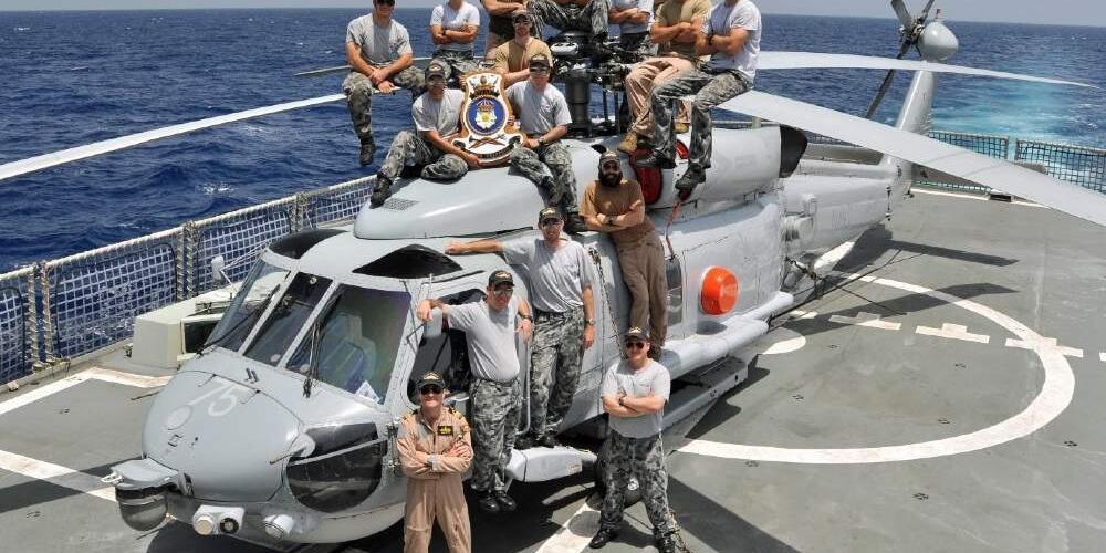 Group of military personal posing on and around a grey helicopter on the back of a ship with the ocean in the background.