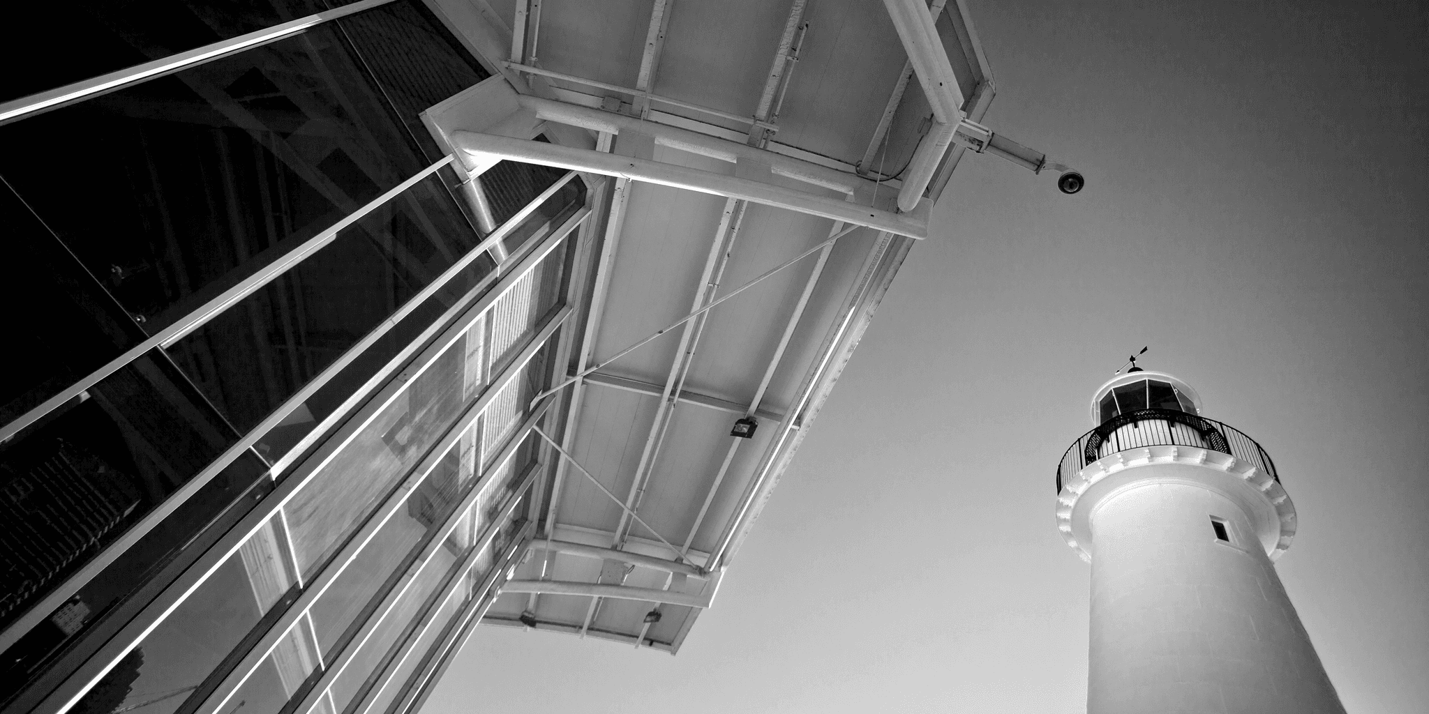 Black and white drematic photo looking up at the edge of a modern building and a lighthouse. 