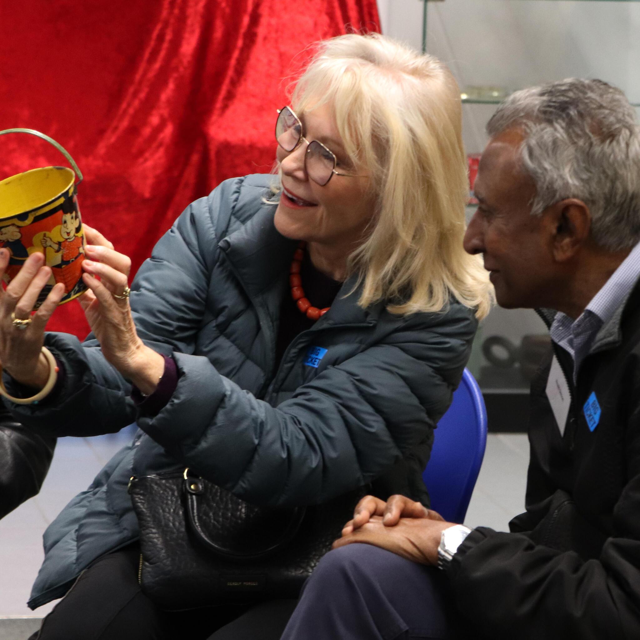 Lady with blonde hair and glasses holding a metal bucket and showing it to a man with dark skin and grey hair.