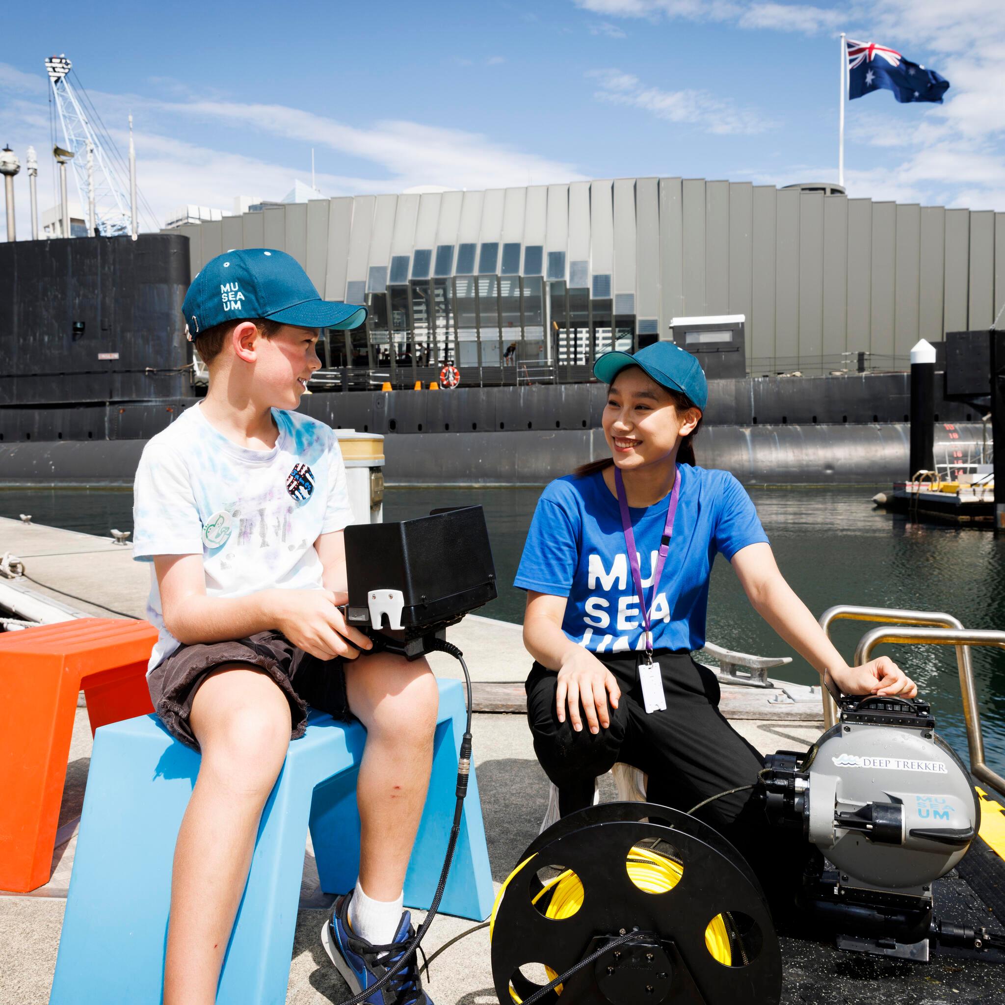 A boy and lady wearing hats and sitting on a wharf. the boy is holding a remote controller. there is a submarine in the background.