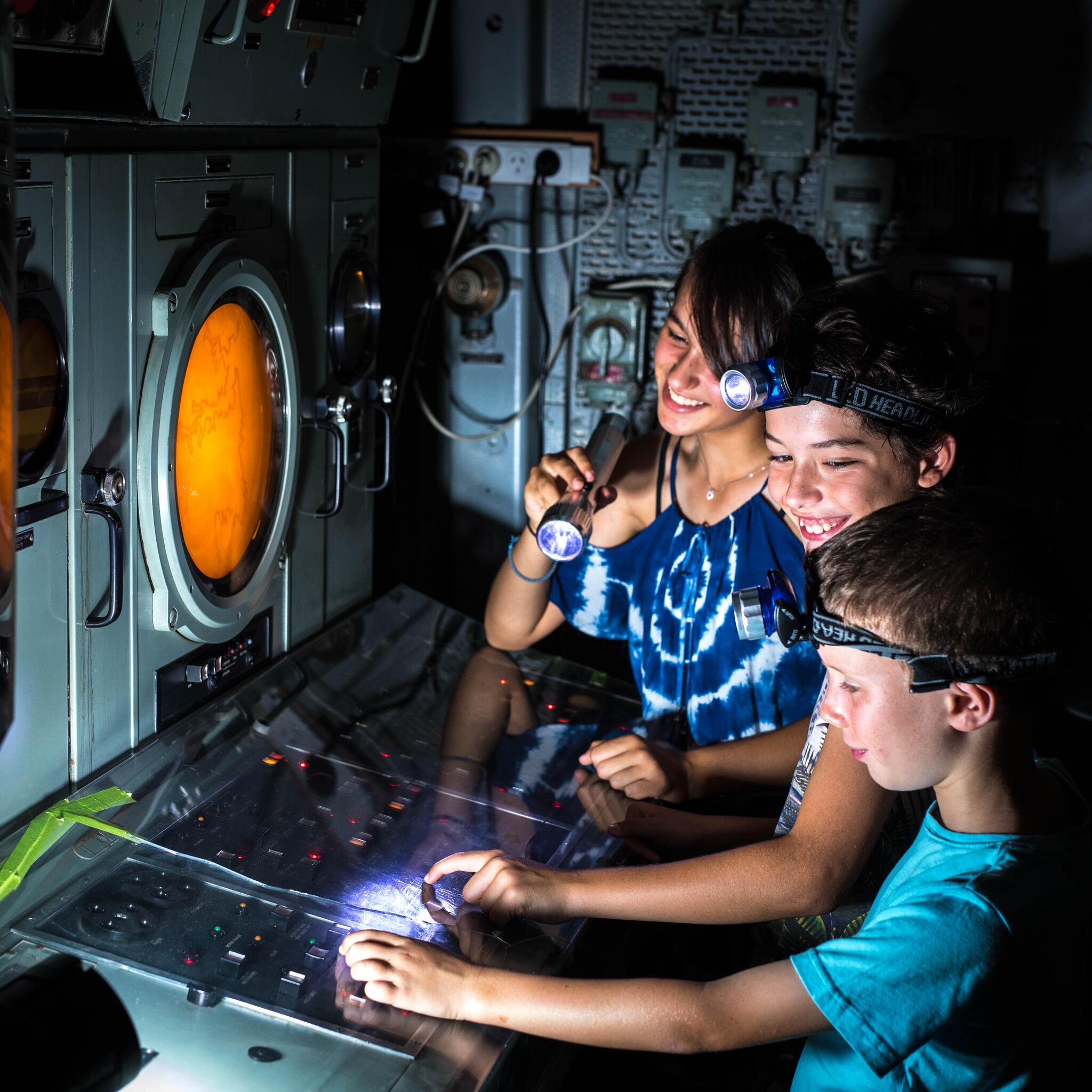 Photo of 3 children in a dark space with torches looking at a piece of technical equipment with a yellow round screen and buttons. 