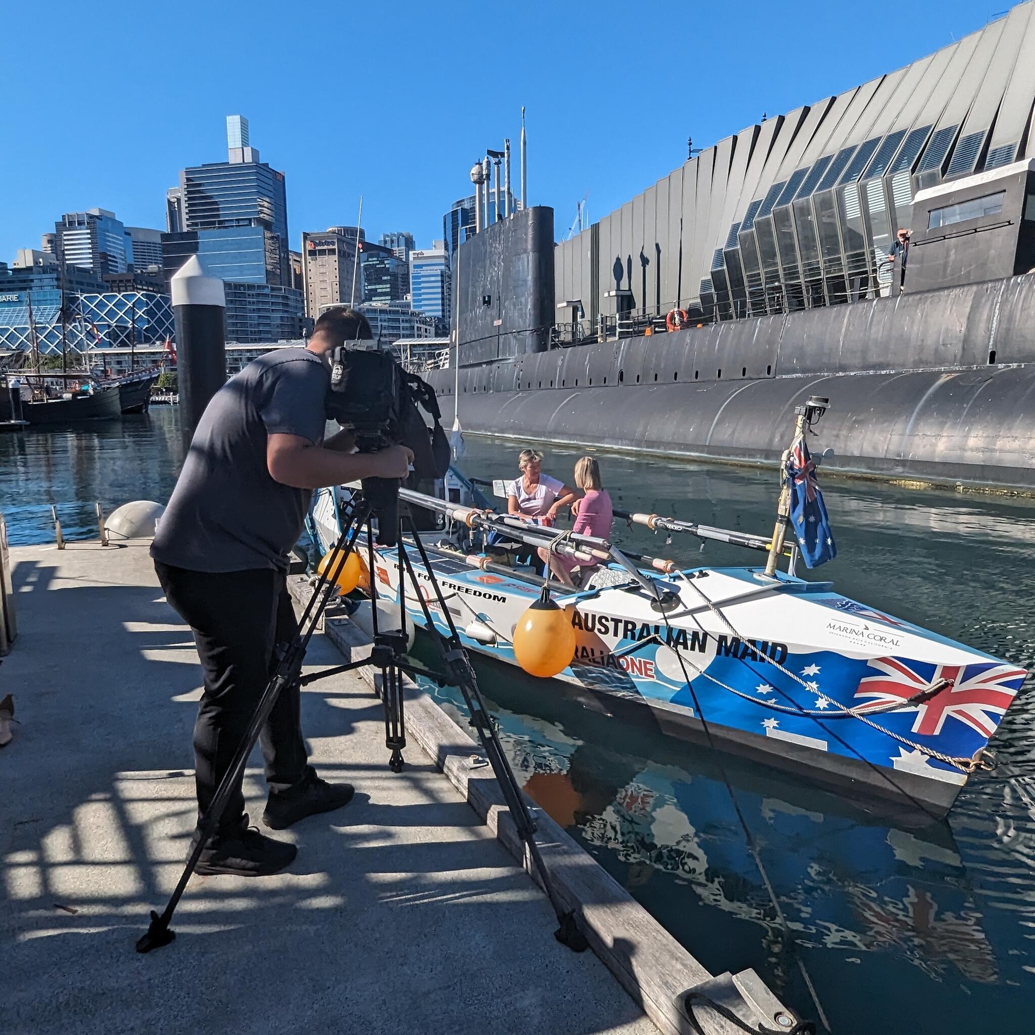 photo showing behind the scenes, with a camera man standing on the wharf, taking video of two ladies in a boat.