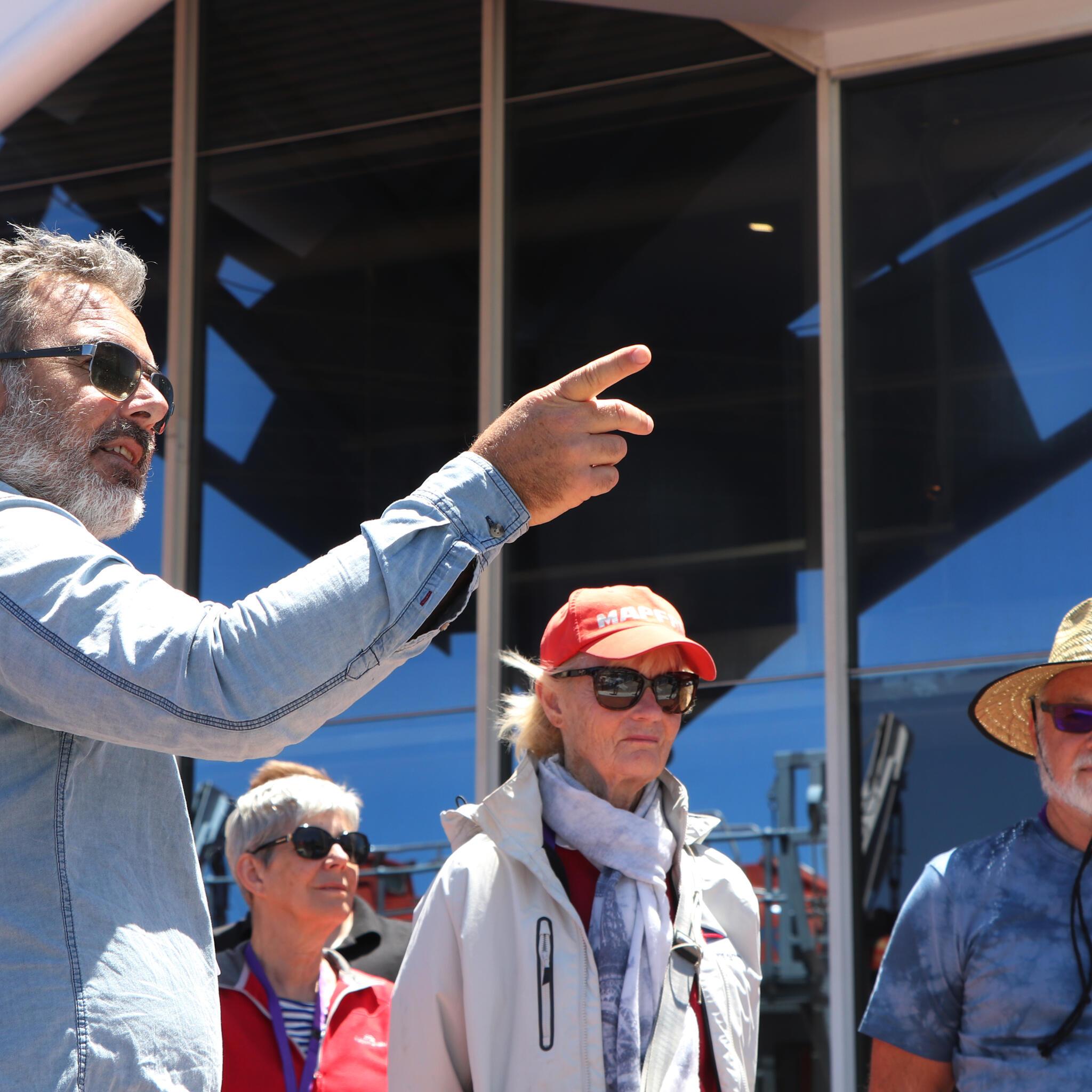 A man with grey hair and his back to the camera is pointing to the right. There are other people in the background wearing hats and sunglasses. 