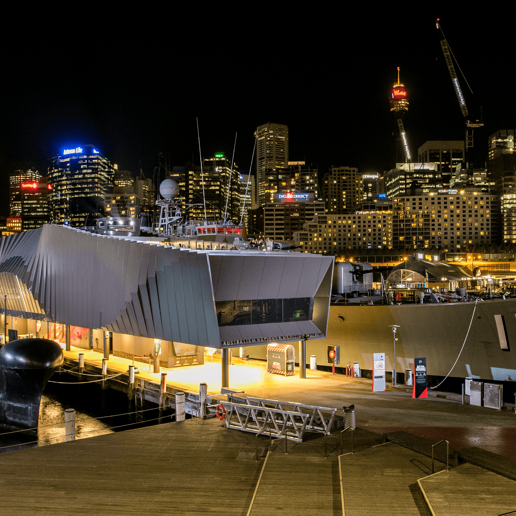 Exterior views of the Waterfront Pavilion taken at night, with the submarine HMAS ONSLOW moored at left and HMAS VAMPIRE on the right. Illuminated city buildings appear in the background.