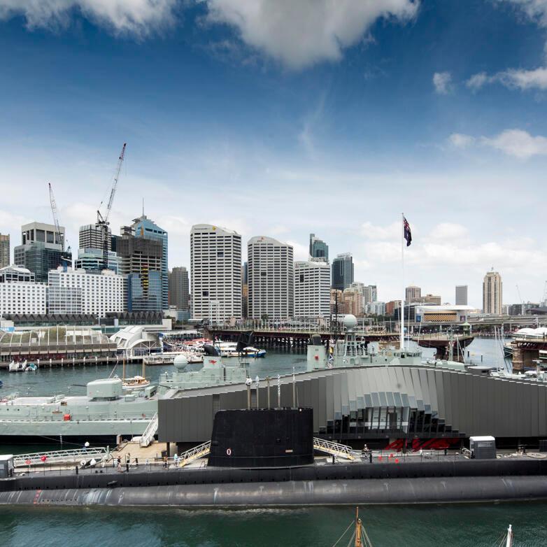 This wide elevated exterior view showcases both the building's unique architecture as well as its location in relation to the city and Darling Harbour precinct. The ONSLOW submarine, shown in profile, features prominently in front of the building. The Pyrmont Bridge and city skyline appear in the background. 