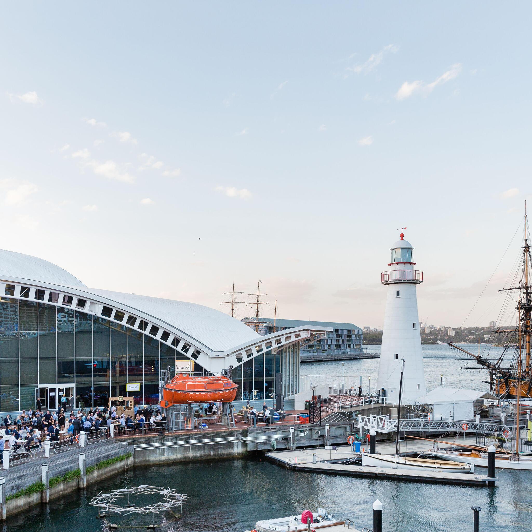 The museum precinct filled with crowds during a corporate event. museum building on the left, with lighthouse and tall ship on the right.