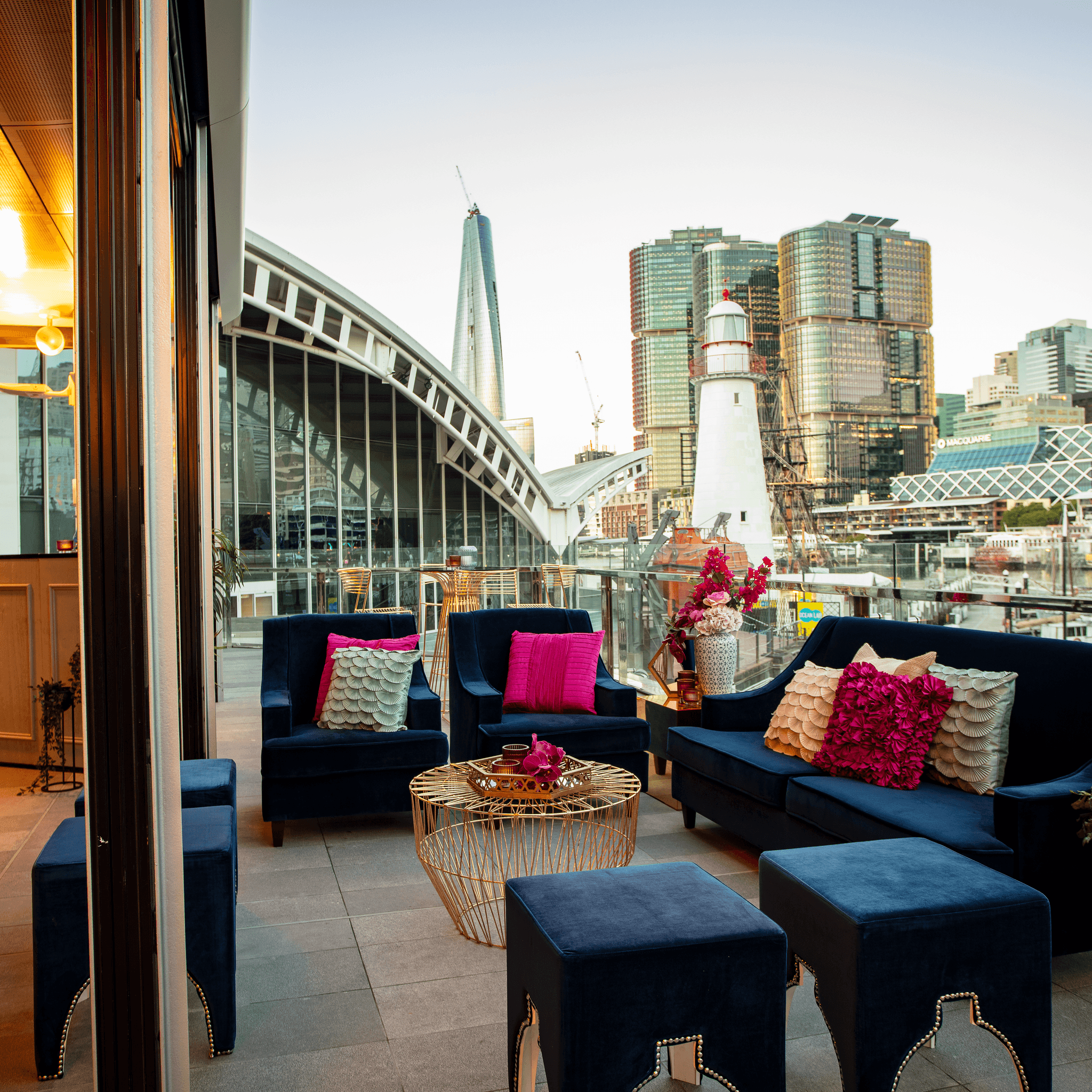 photo of outdoor balcony with blue event furniture and a lighthouse and Sydney skyline in the background. 