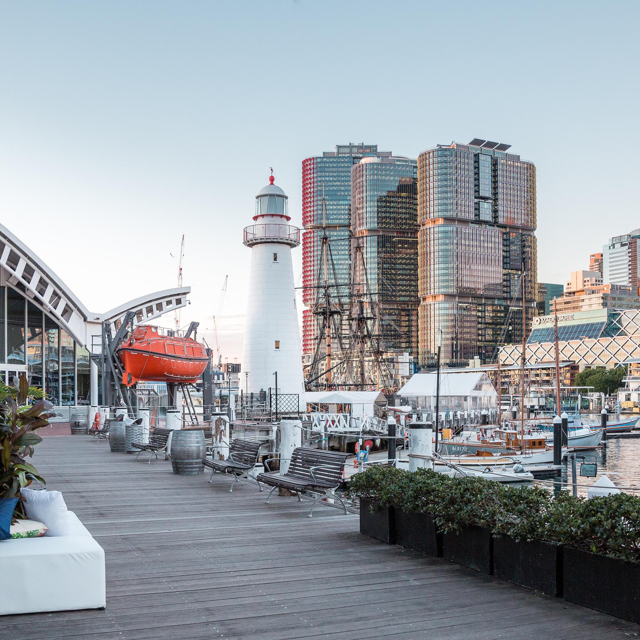 photo of outside the museum, with the boardwalk in the foreground, and a lighthouse and the Sydney skyline in the background on a sunny day. 