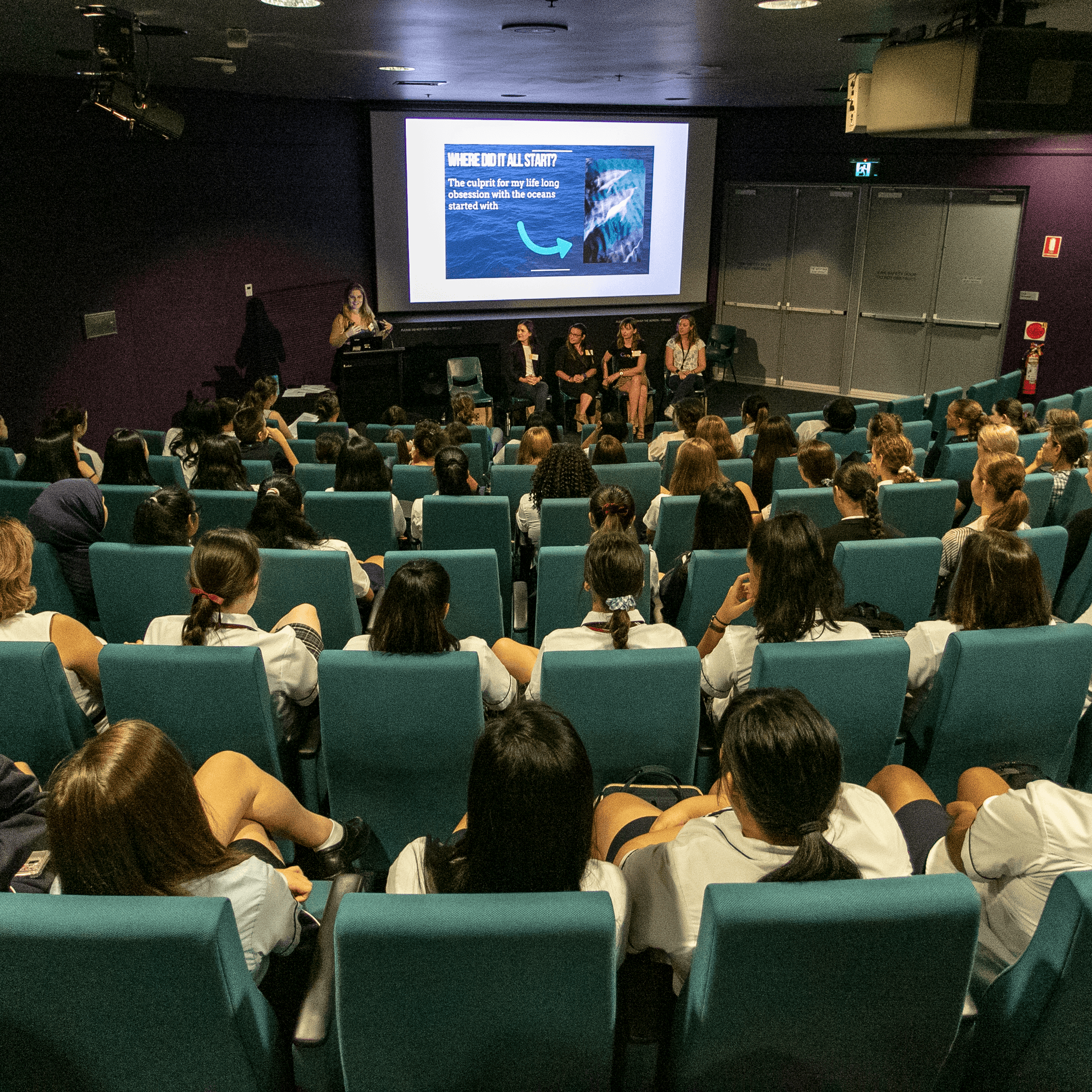 Photo from the back of the theatre, with school students watching a speaker and presentation at the front of the room.