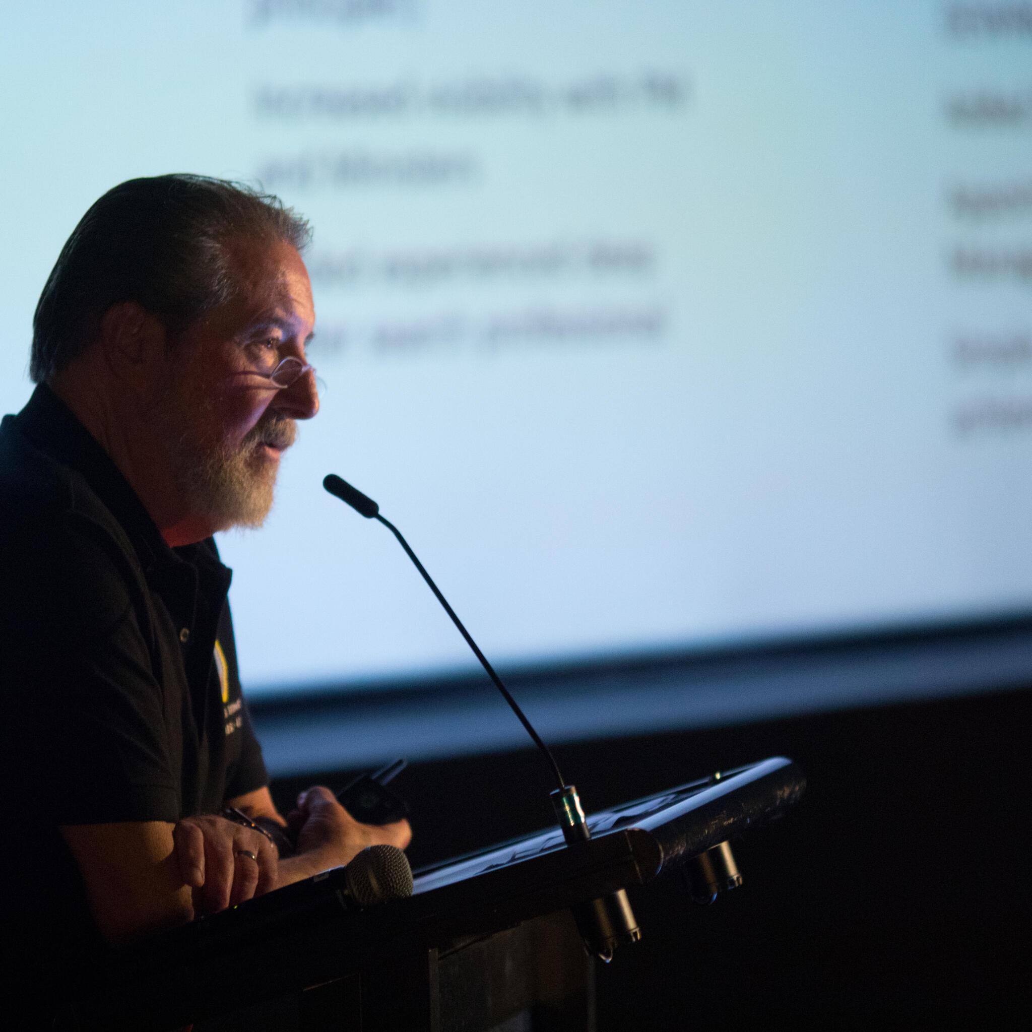 Photo of a side view of a male speaker taking into a lectern microphone, with a presentation in the background.