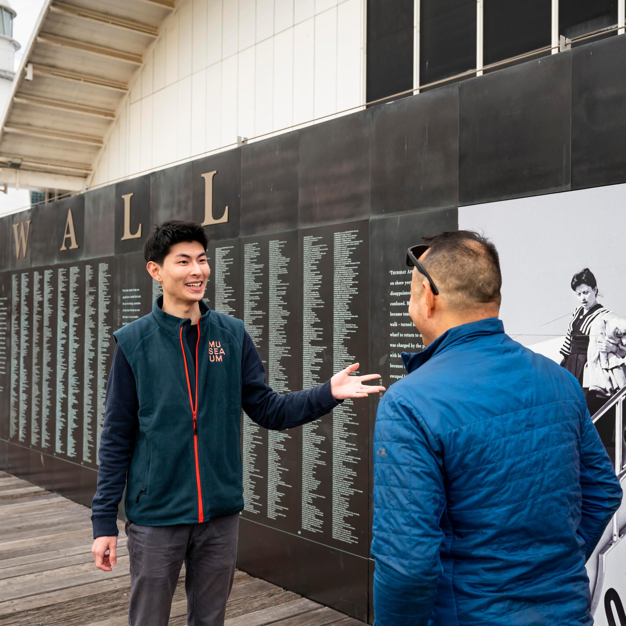 Male volunteer in a uniform showing a visitor names engraved on the national monument to migration