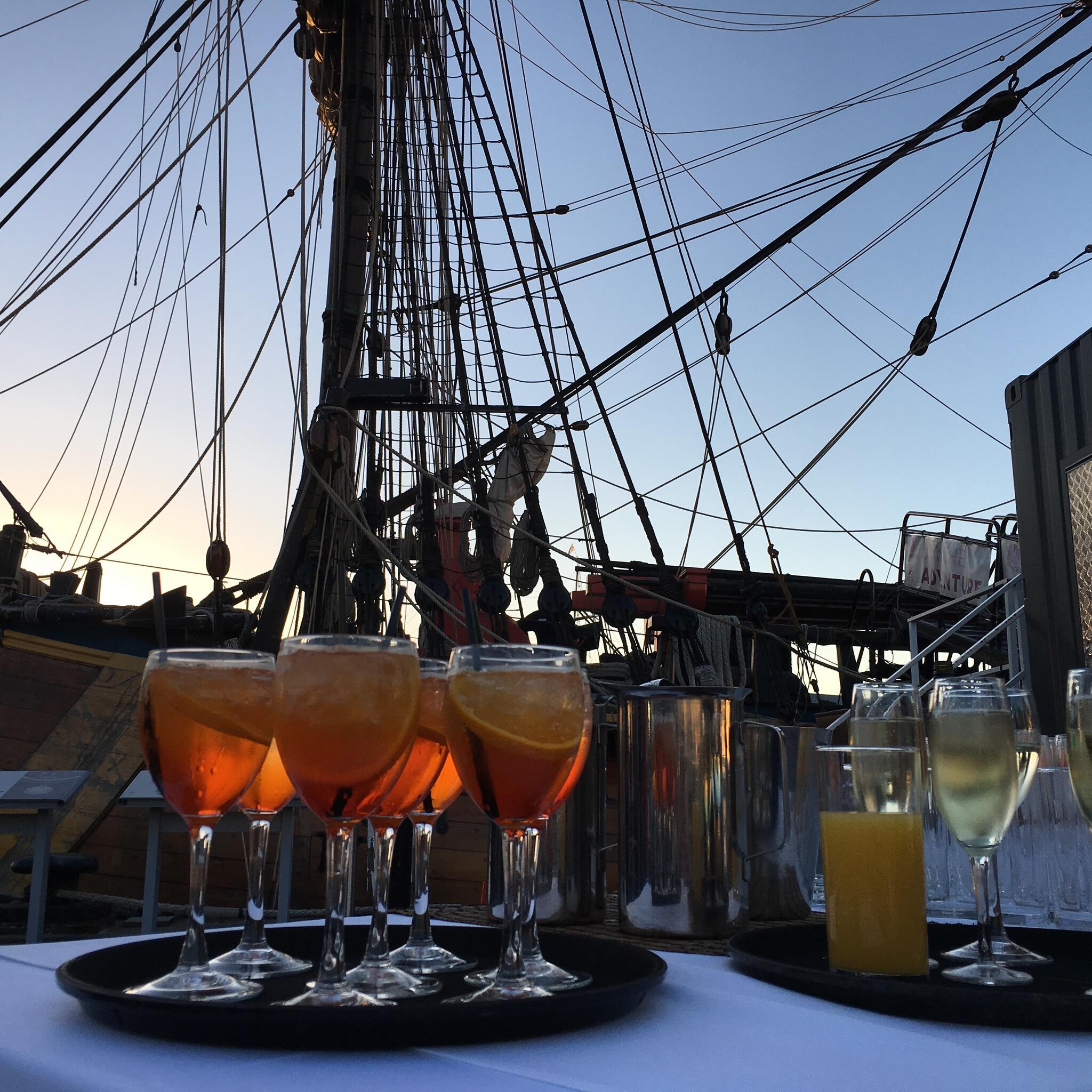 Photo of cocktails on a table in front of tall ship masts. 