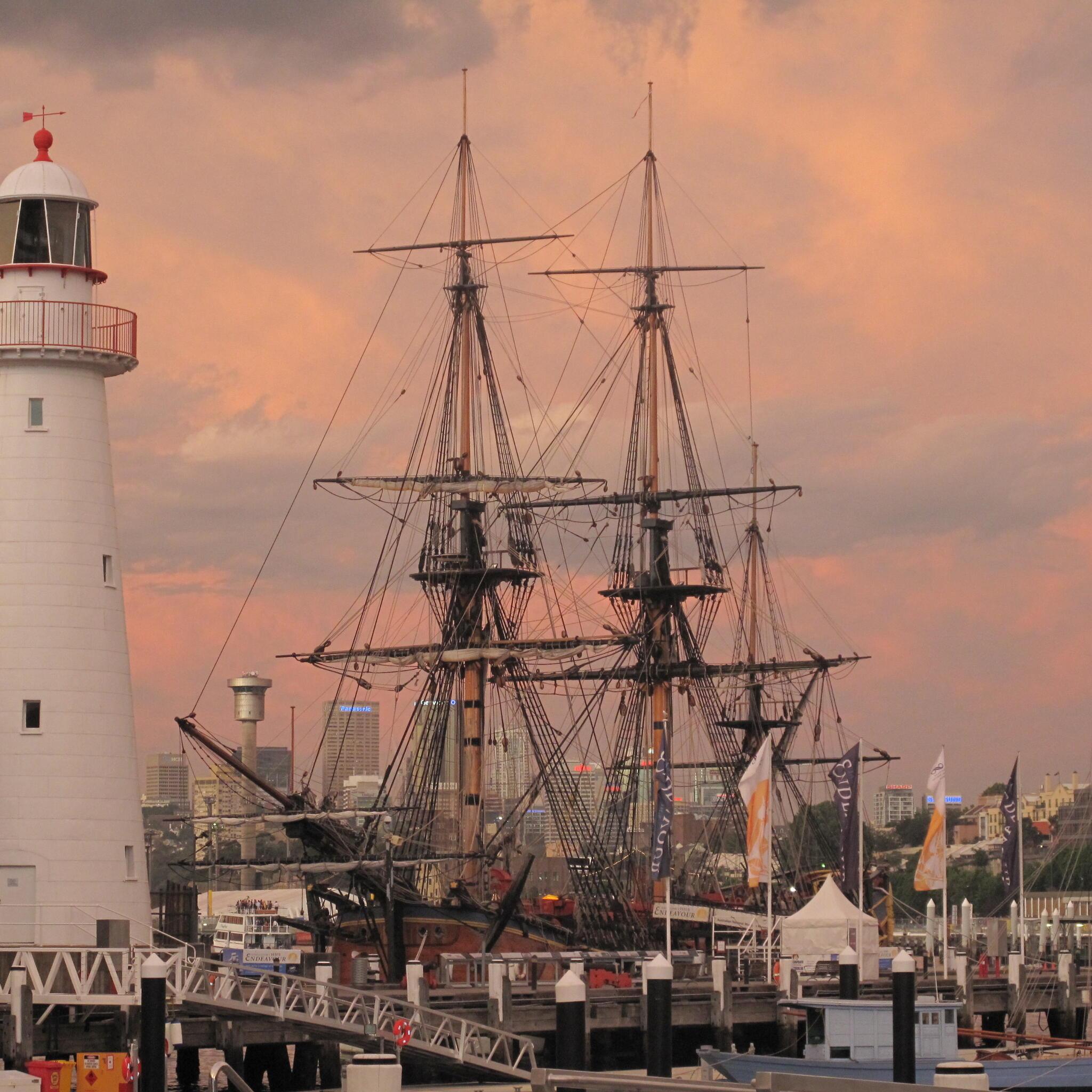 Photo of tall ship and lighthouse, with a pink orange sunset. 