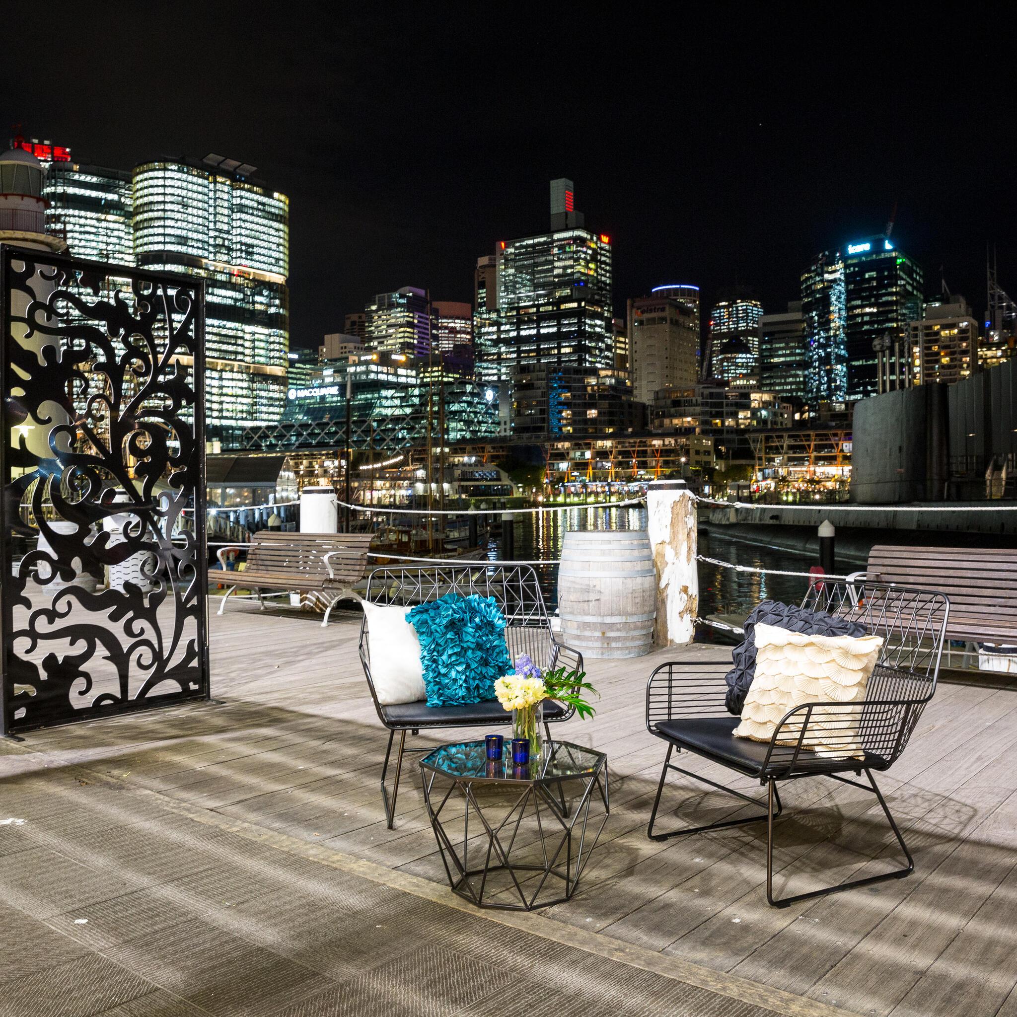 photo taken at night of boardwalk with outdoor furniture and Sydney skyline in the background. 