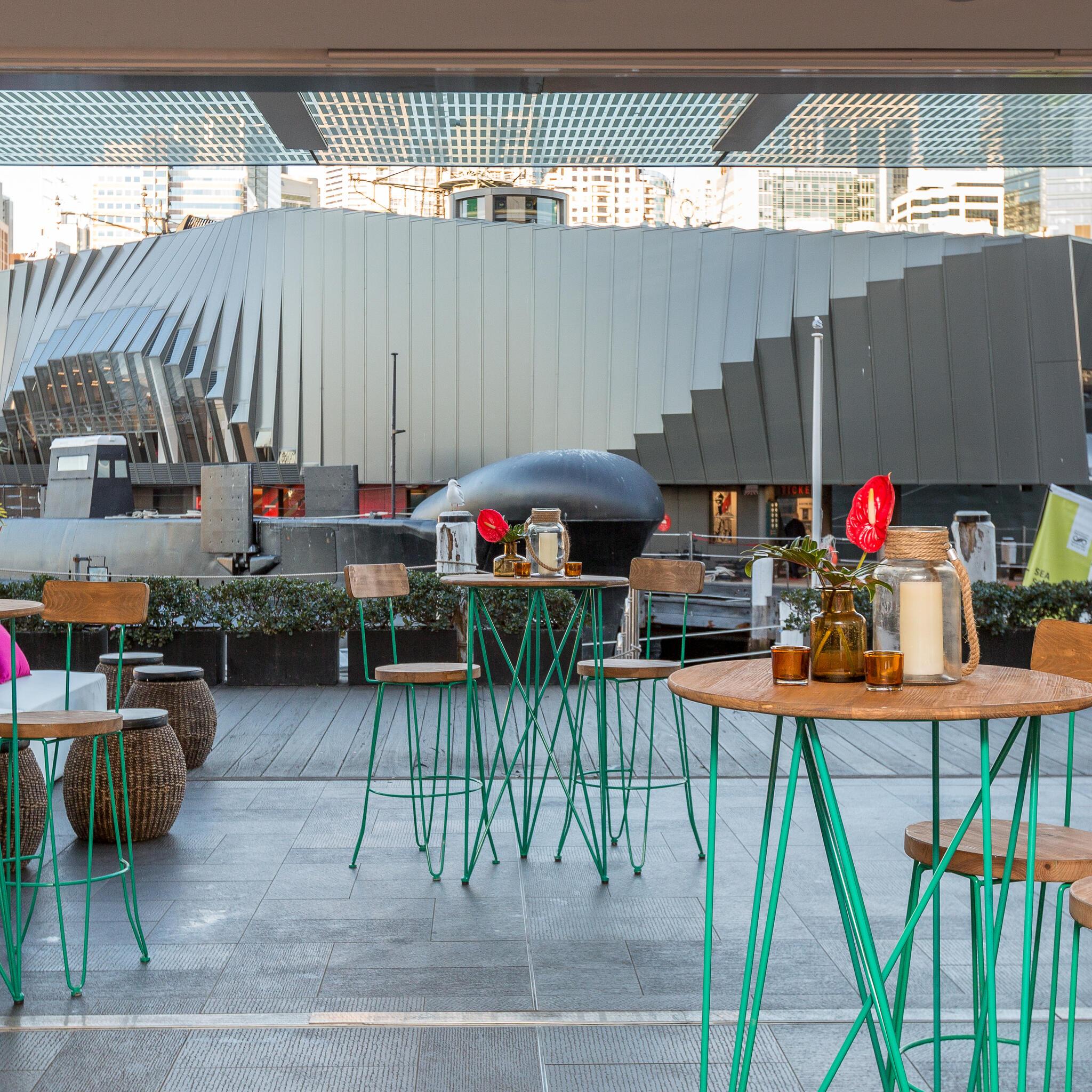 Photo from an interior opening into an exterior space, with natural and teal coloured event furniture and a submarine in the background. 