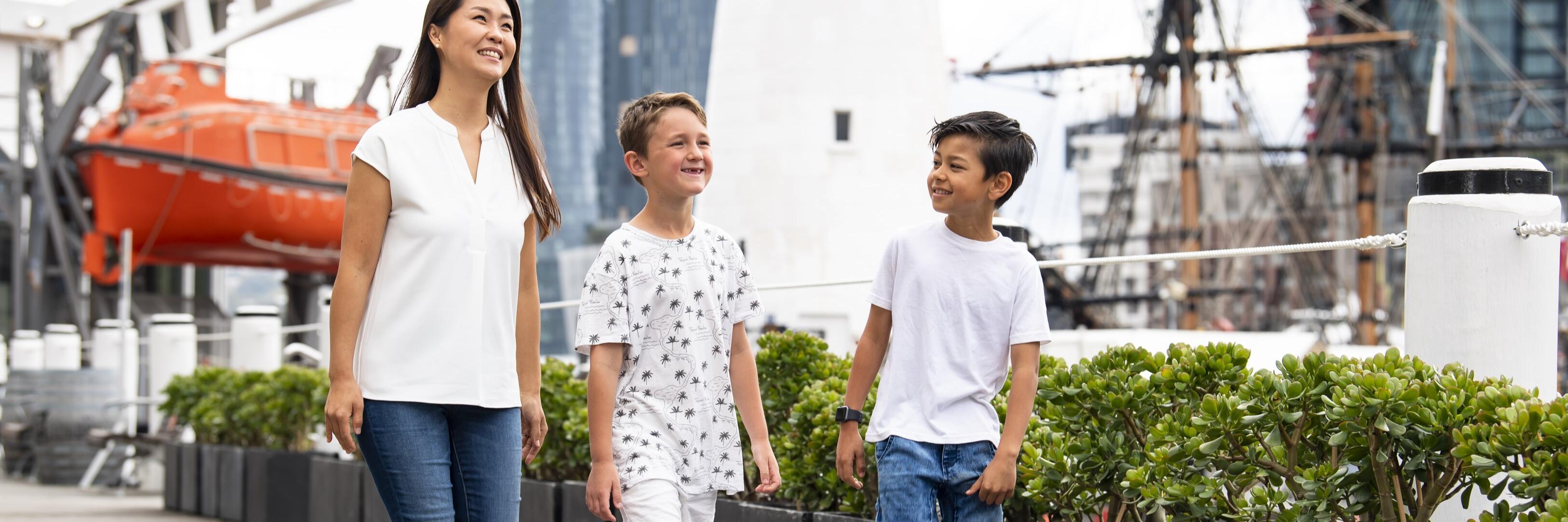 Photo of a women and two boys walking down a boardwalk, with shrubs, a lighthouse, tall ship masts and orange lifeboat in the background. 