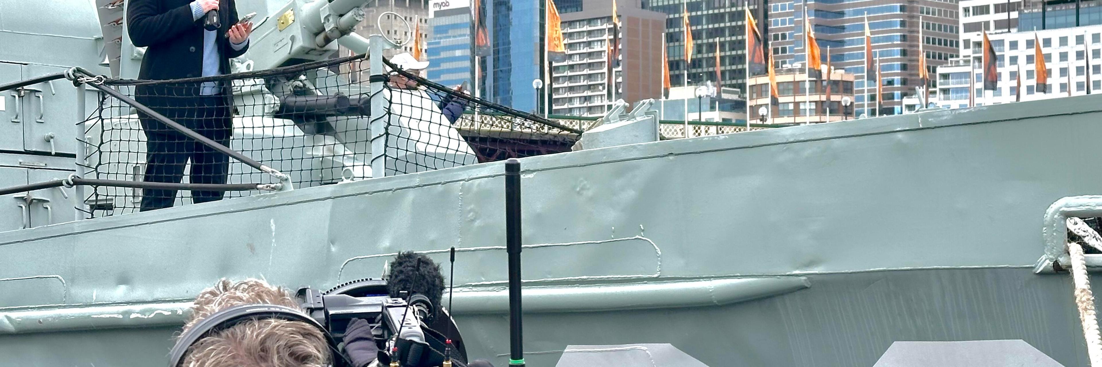 Photo showing behind the scenes off a TV shoot on a navy vessel, with a camera man in the foreground, and a male presenter standing on the boat behind.